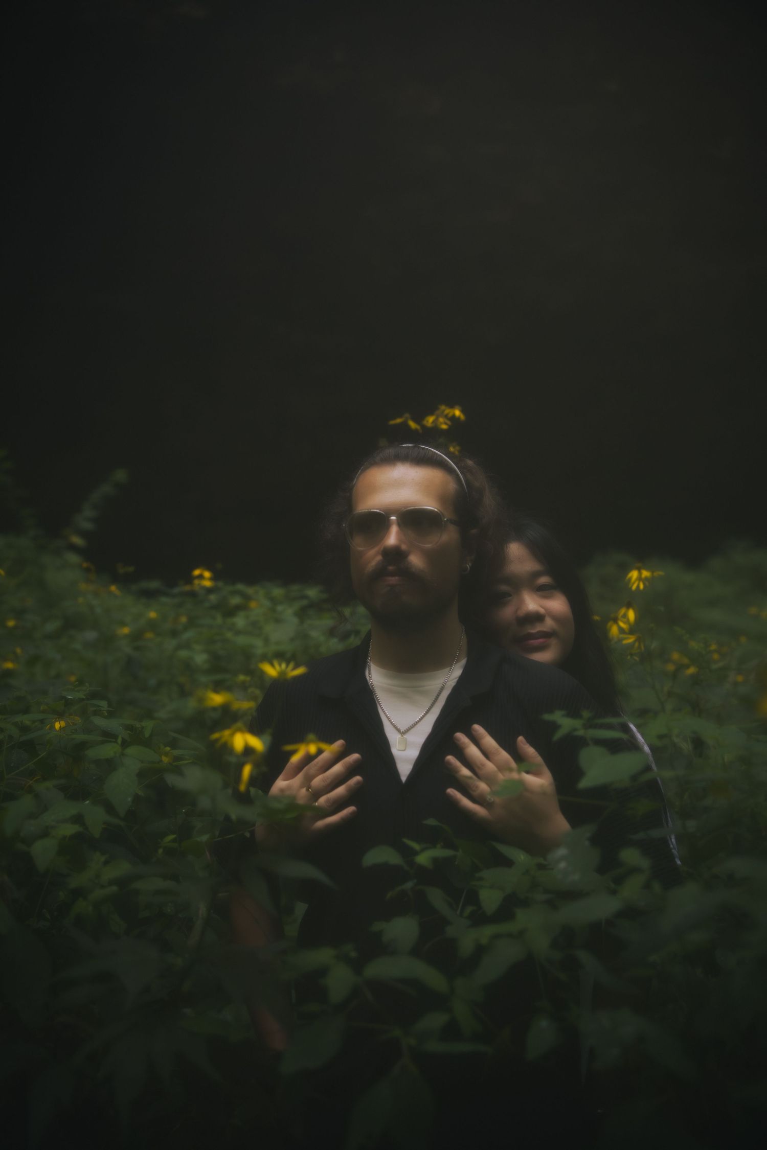 Moody portrait of two people standing among flowers in a dark garden setting, creating a dramatic and mysterious atmosphere.