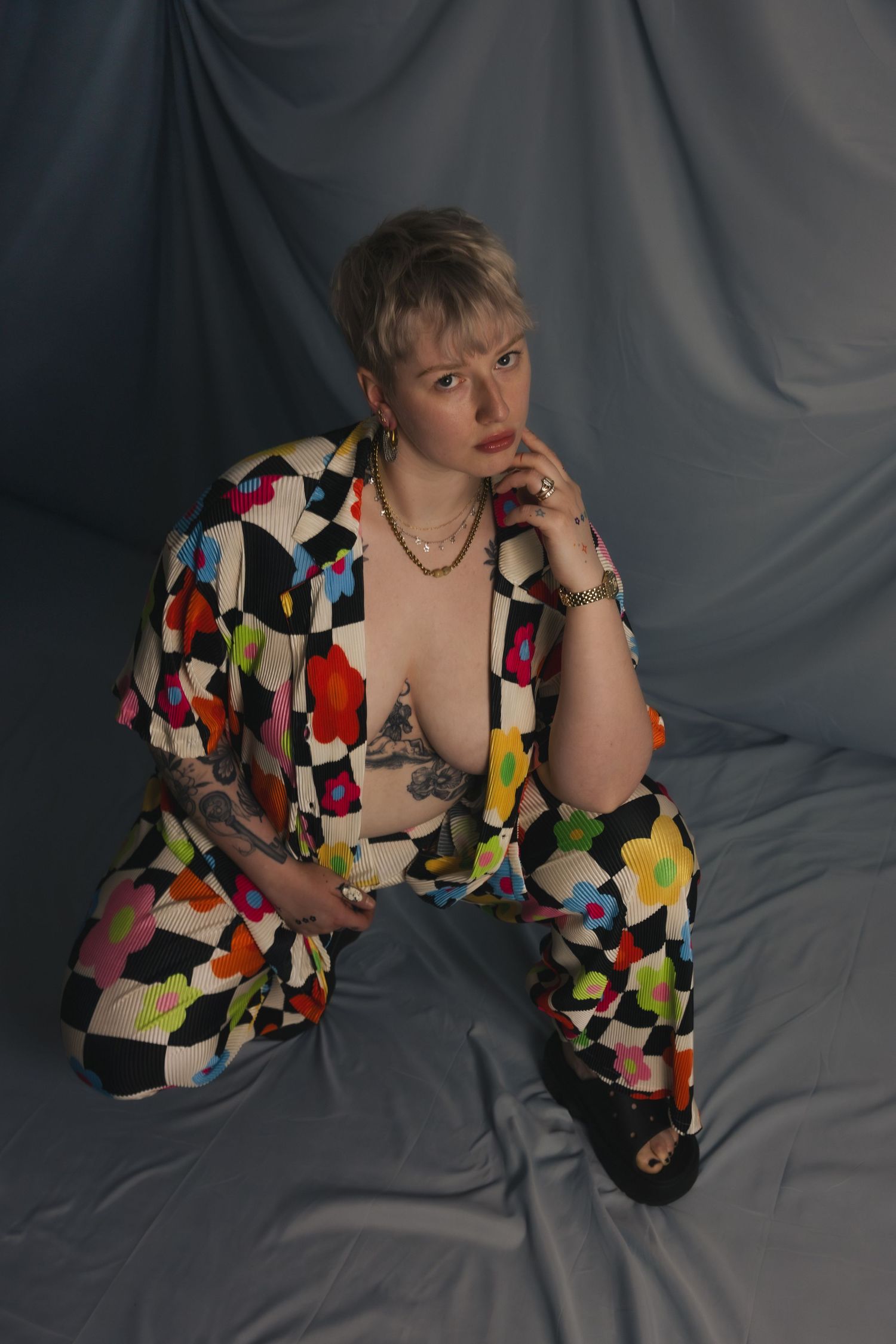 Person in colorful geometric floral print suit seated against gray draped fabric backdrop.