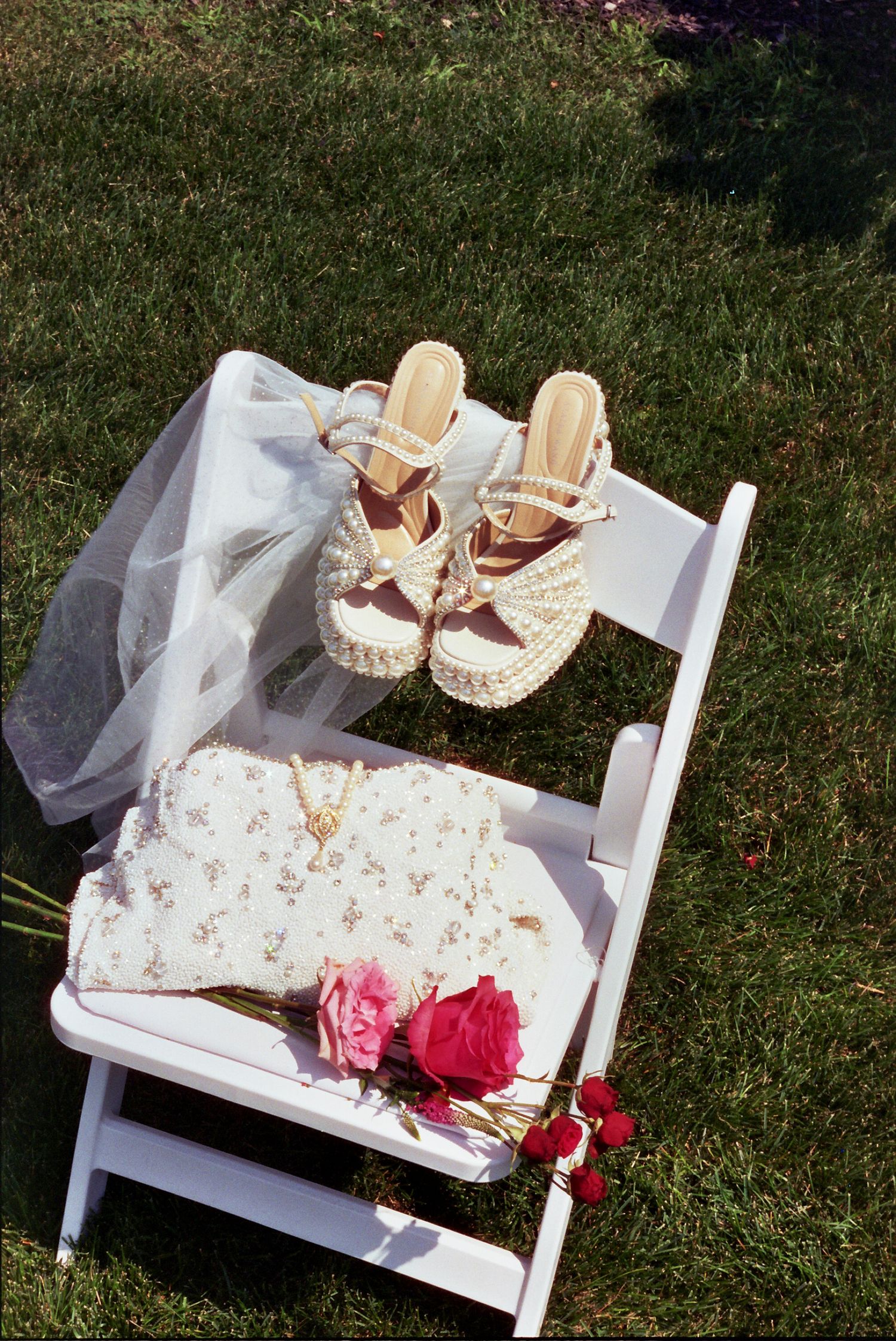 Summer wedding accessories displayed on white chair with pink flowers, floral dress, heeled sandals and veil on grass.