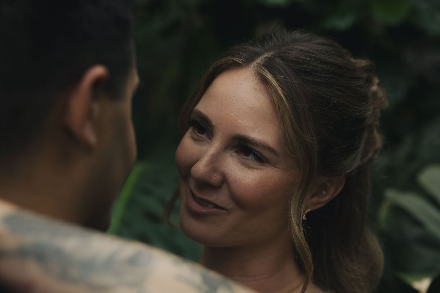 A smiling woman with an updo hairstyle gazes at a tattooed man amid lush green tropical foliage.