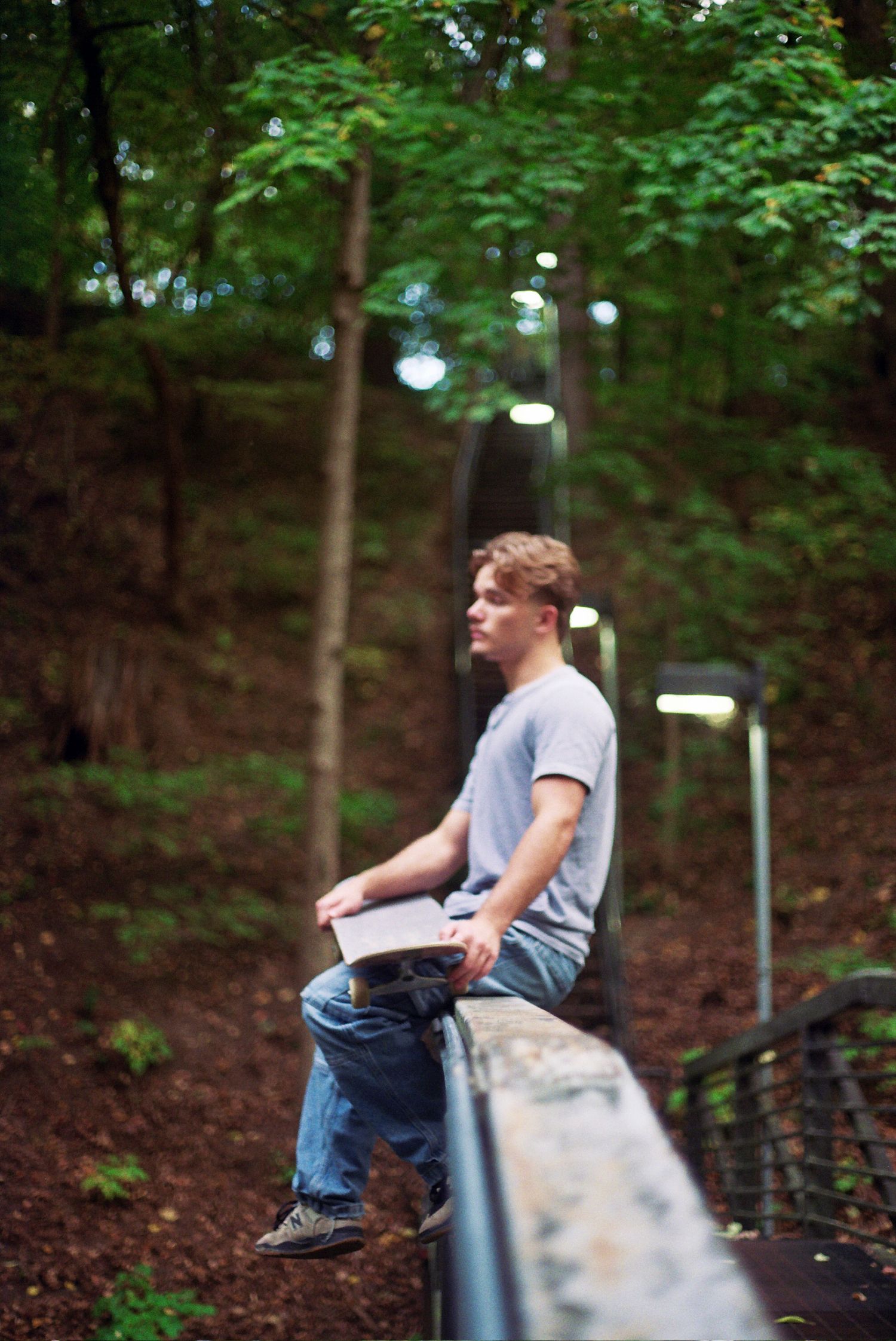 Young man in gray t-shirt and jeans sitting on a stone railing holding a book in a wooded park setting.