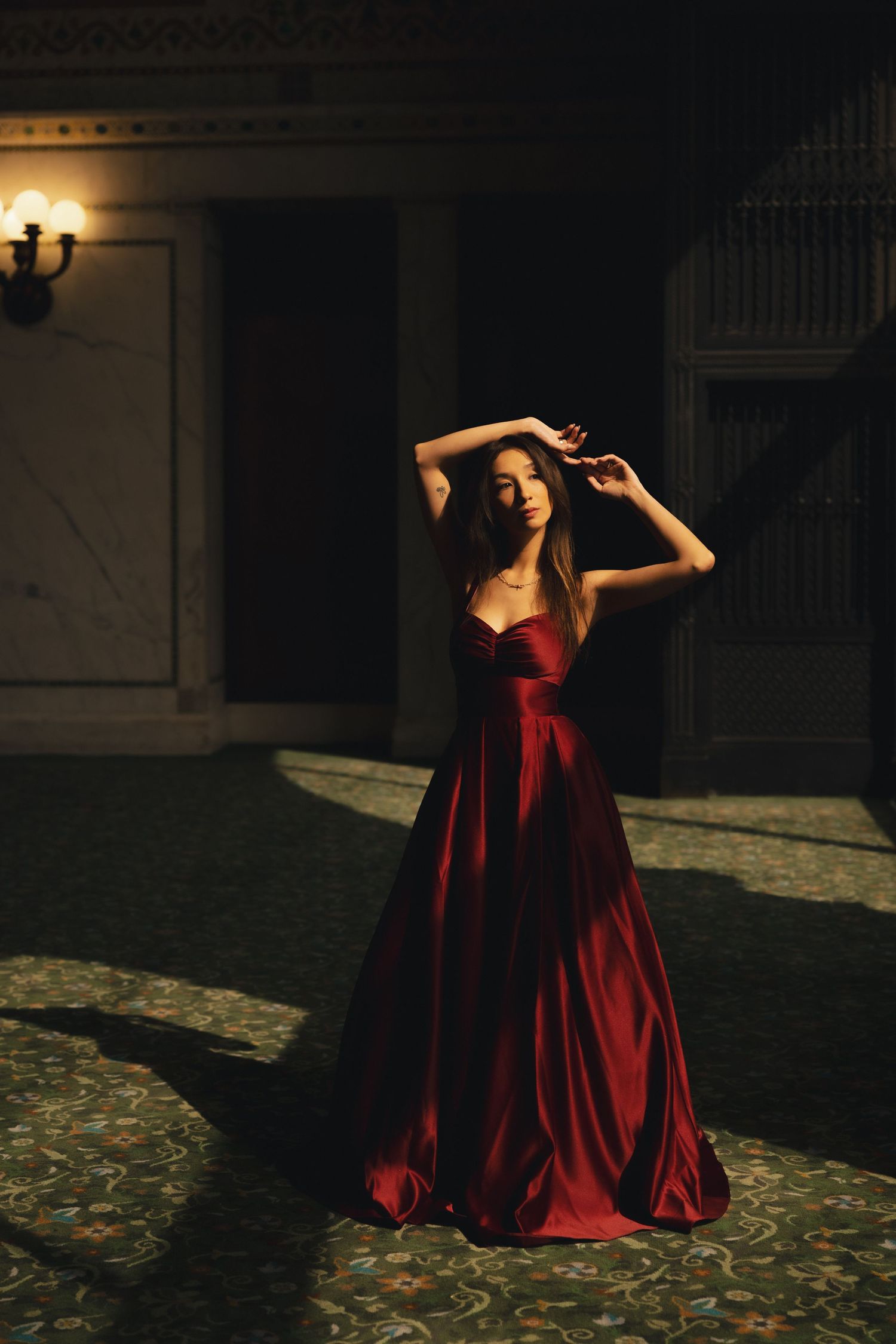 Woman in elegant red satin ball gown posing gracefully in a dimly lit ornate hall.