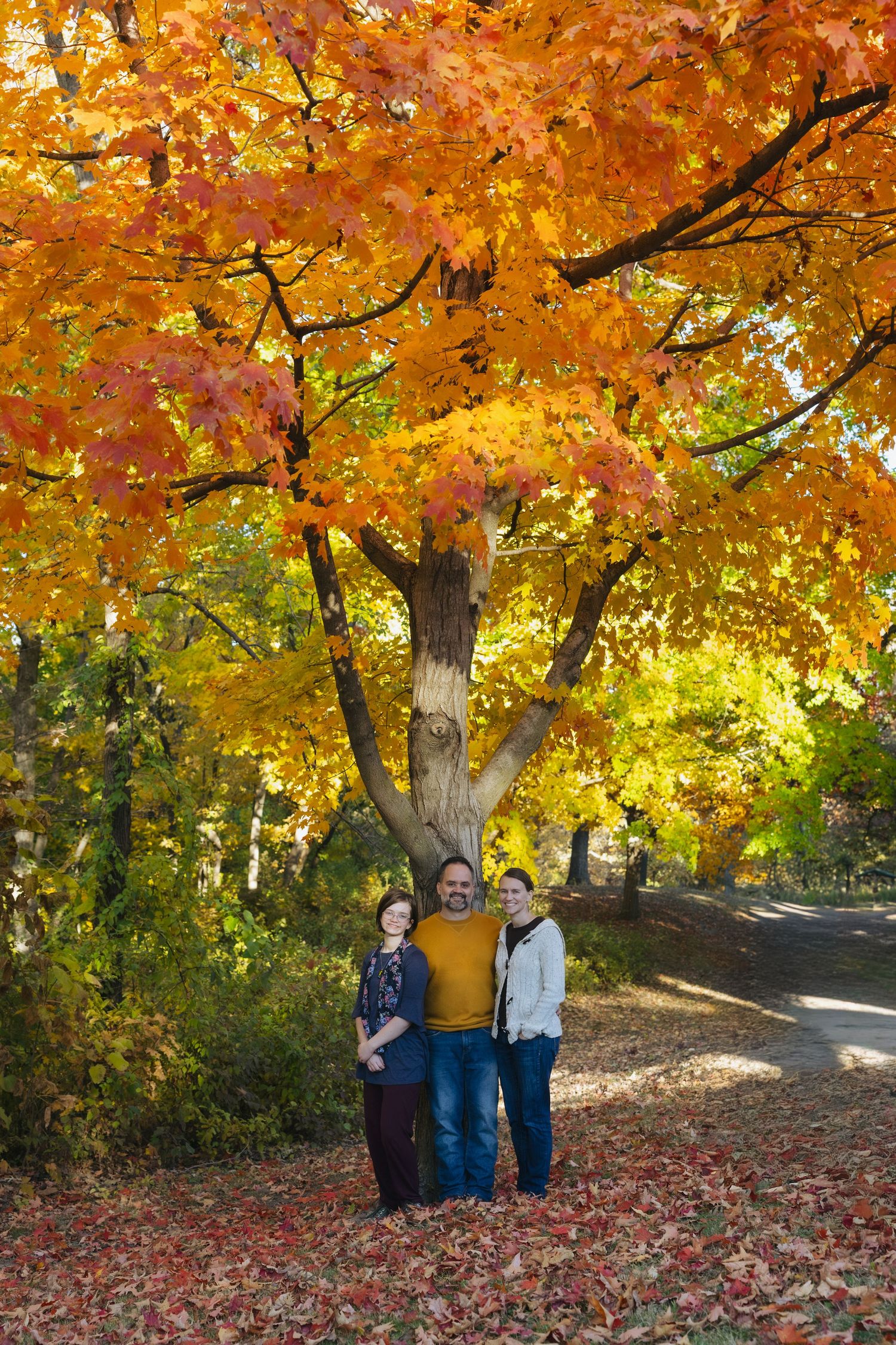 Three people pose under a vibrant autumn maple tree with orange and yellow leaves on a sunny fall day.