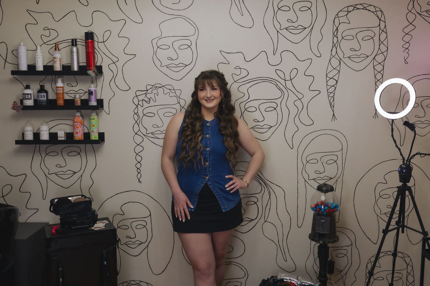 Woman smiling in a hair salon, standing against a wall mural with ring light and product shelves nearby.