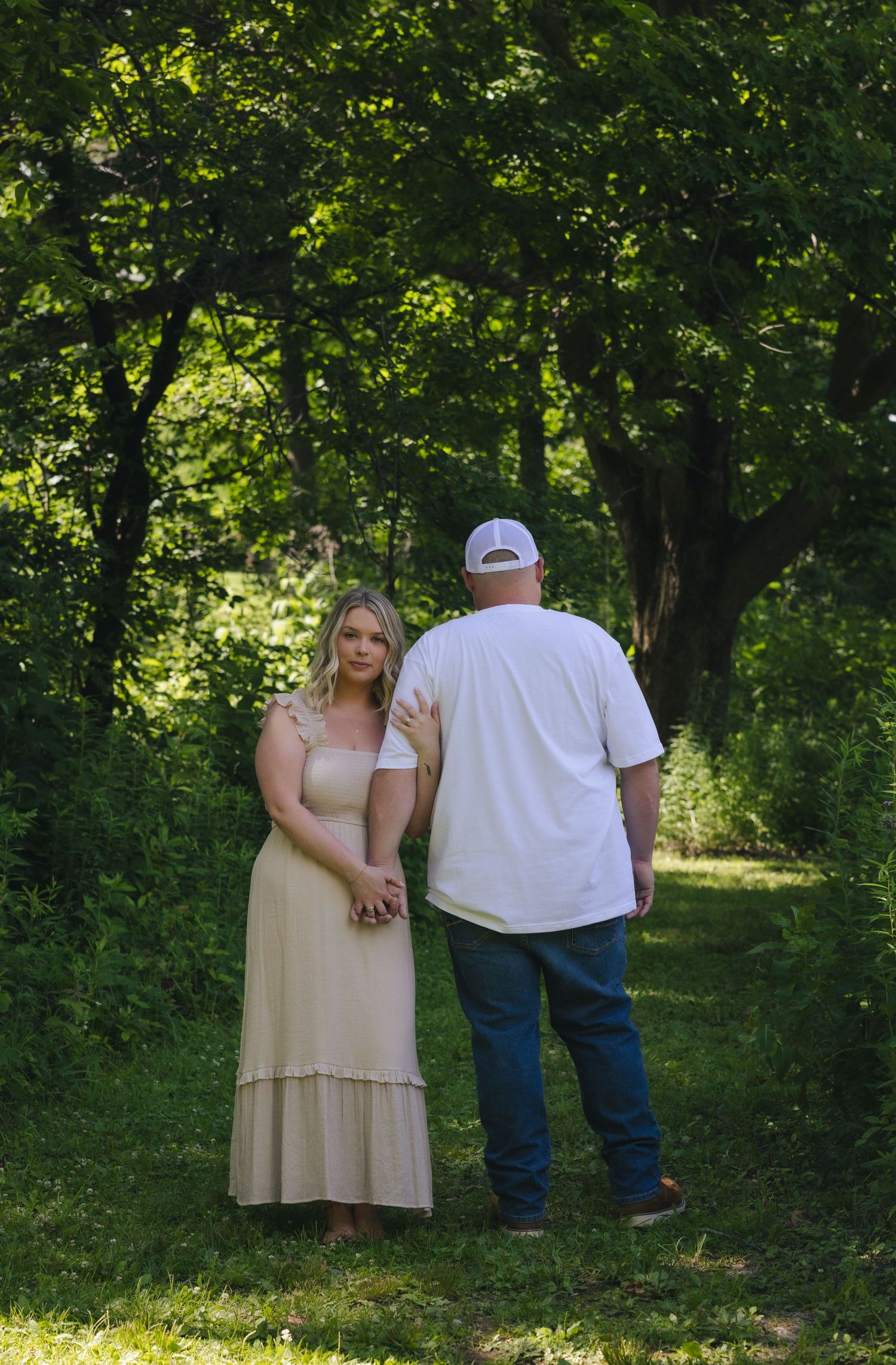 Couple holding hands on a lush green forest path, woman in cream maxi dress, man in white shirt and jeans.
