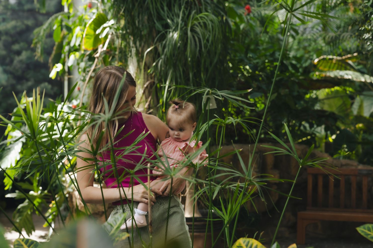 A woman holds a baby among lush tropical greenery in a sunlit botanical garden setting.