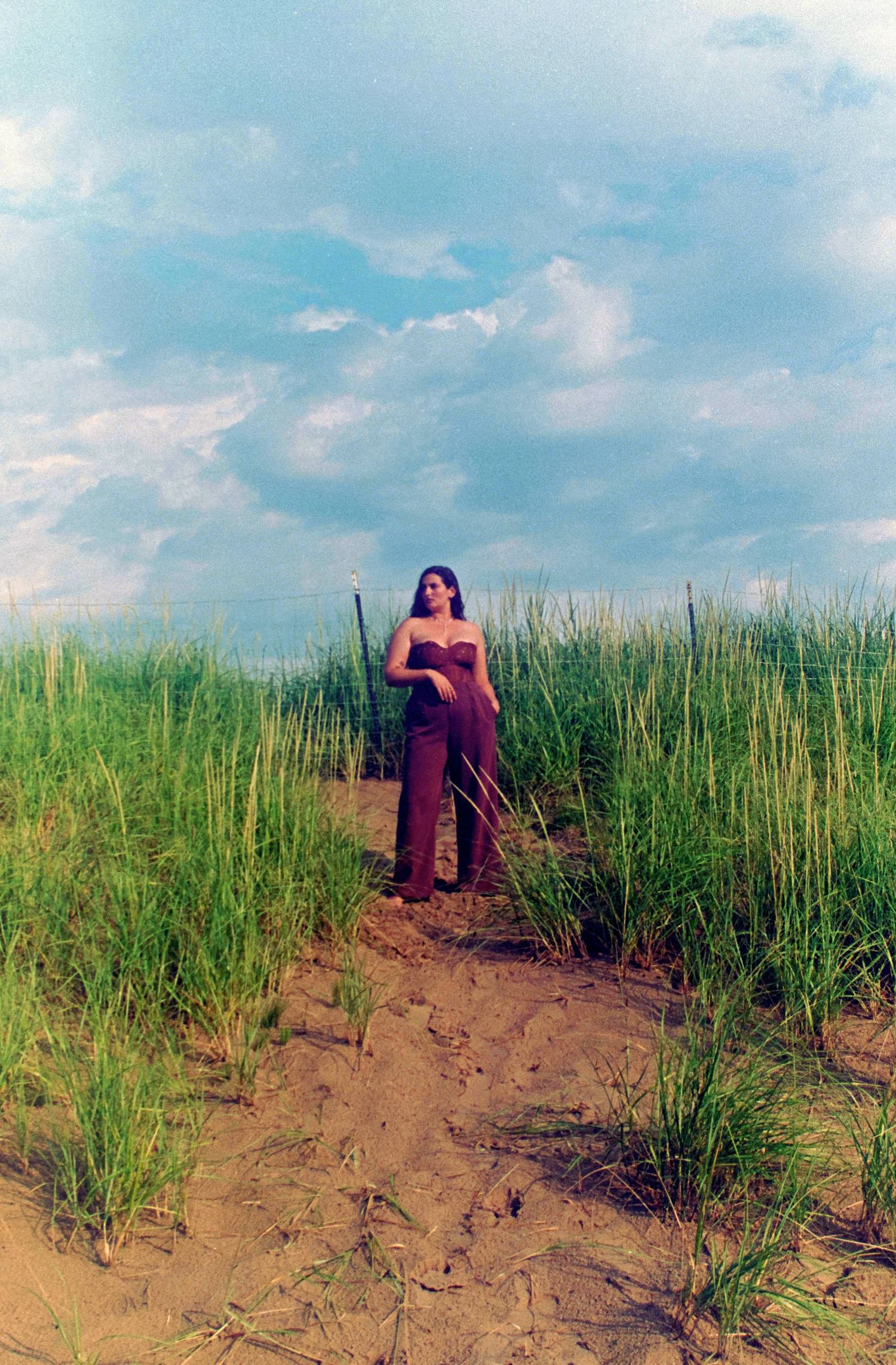 Woman in brown jumpsuit standing on sandy path surrounded by tall green grass under a dramatic cloudy sky.