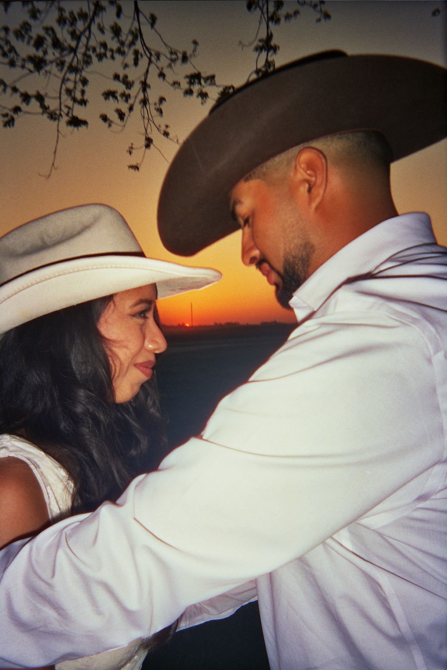 Two people in cowboy hats share an intimate moment against a sunset backdrop while dressed in western attire.