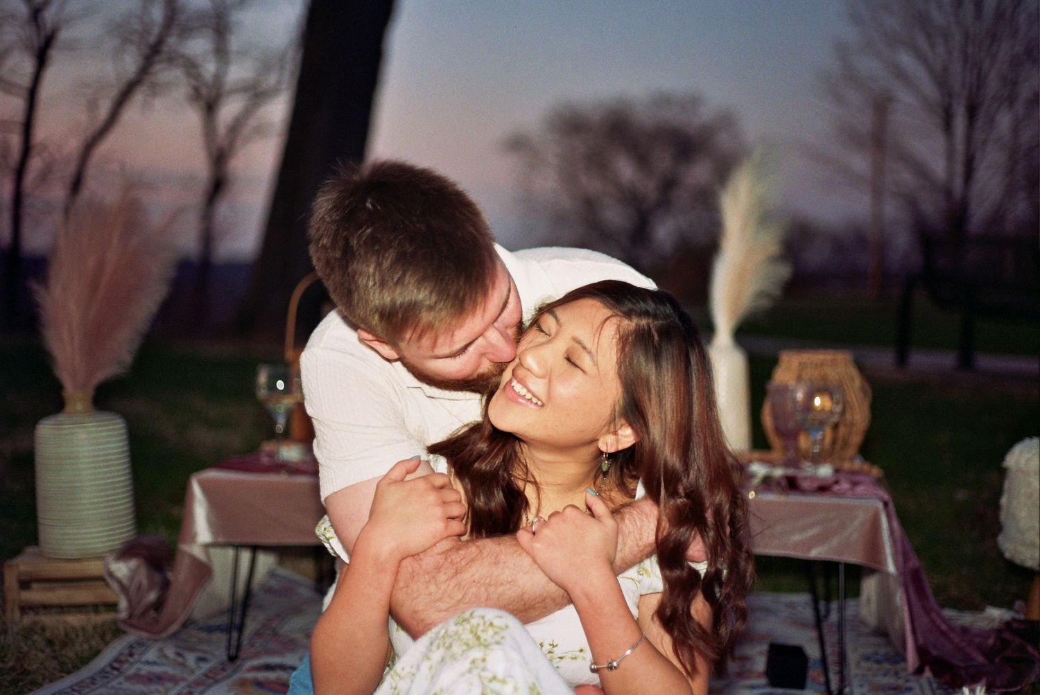 A romantic couple laughs together during an outdoor picnic at sunset with wine glasses and table settings visible.