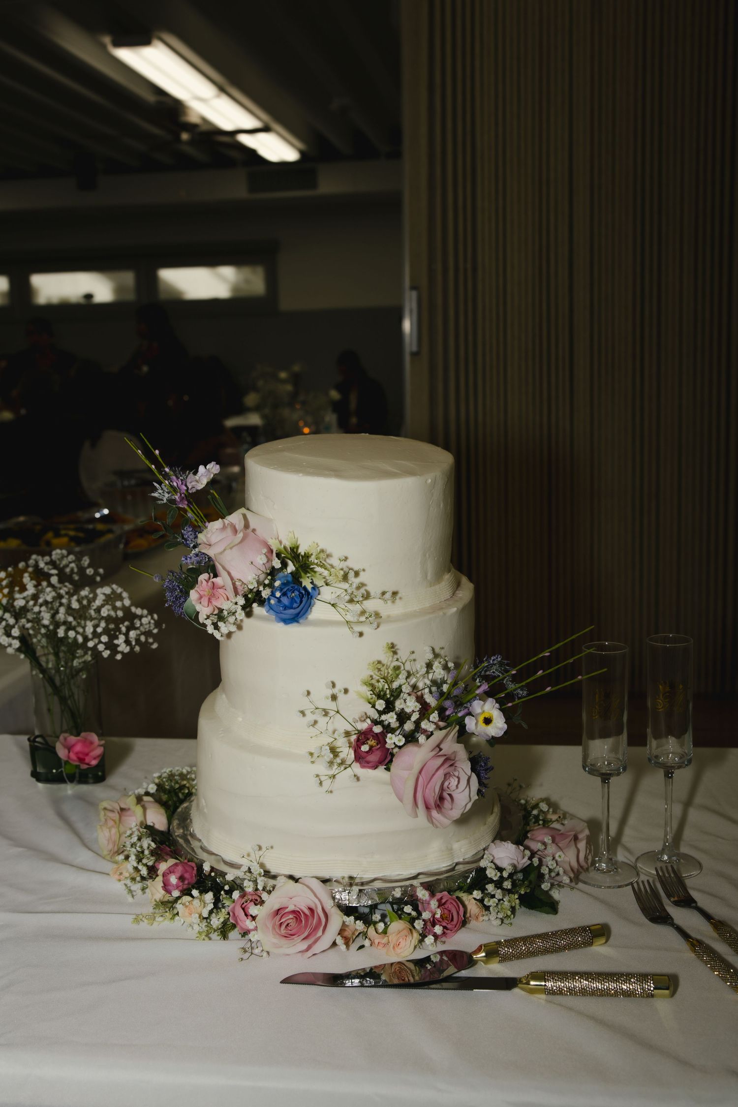 Three-tiered white wedding cake decorated with fresh pink roses, blue flowers and baby's breath on a white tablecloth.