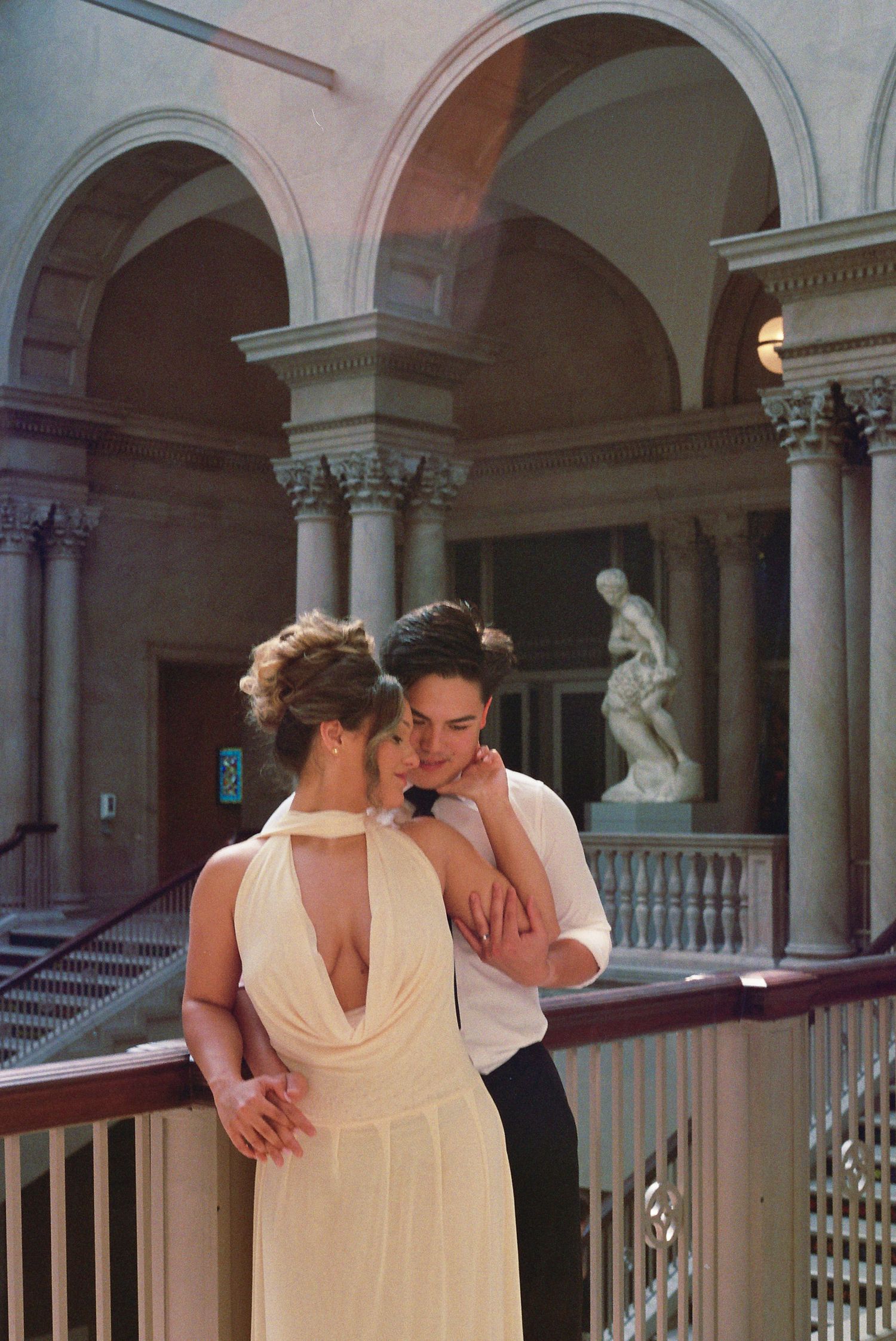 A romantic moment in an elegant museum hallway with arched architecture and classical statuary in the background.