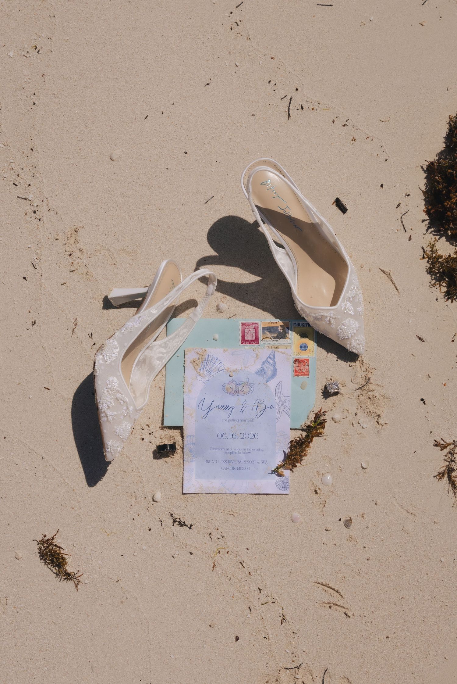 White lace bridal heels and wedding invitation suite laid on sandy beach with colorful stamps and seaglass accents.