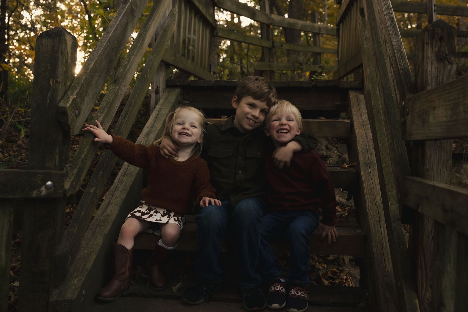 Three children sit together on wooden stairs outdoors during golden hour, smiling and laughing.