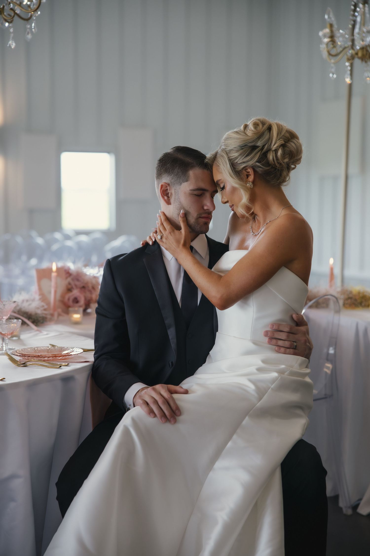 Romantic wedding couple sharing intimate moment at elegant reception with crystal chandeliers and soft lighting.