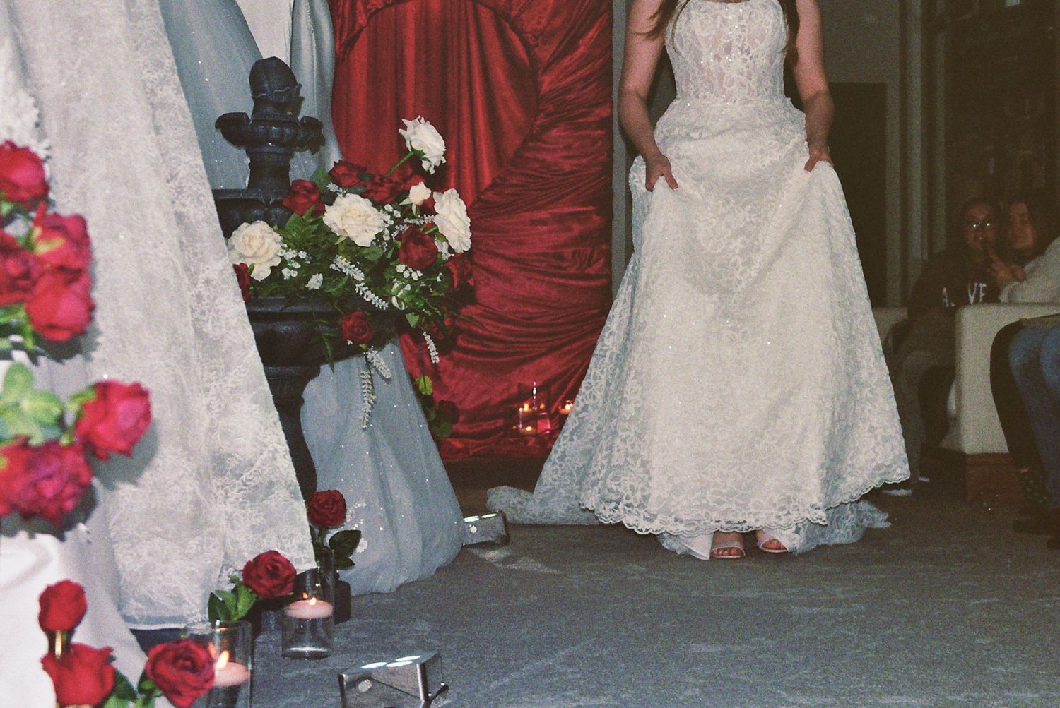 Bride in white lace wedding gown stands near red floral arrangements and draped fabric during a wedding ceremony.
