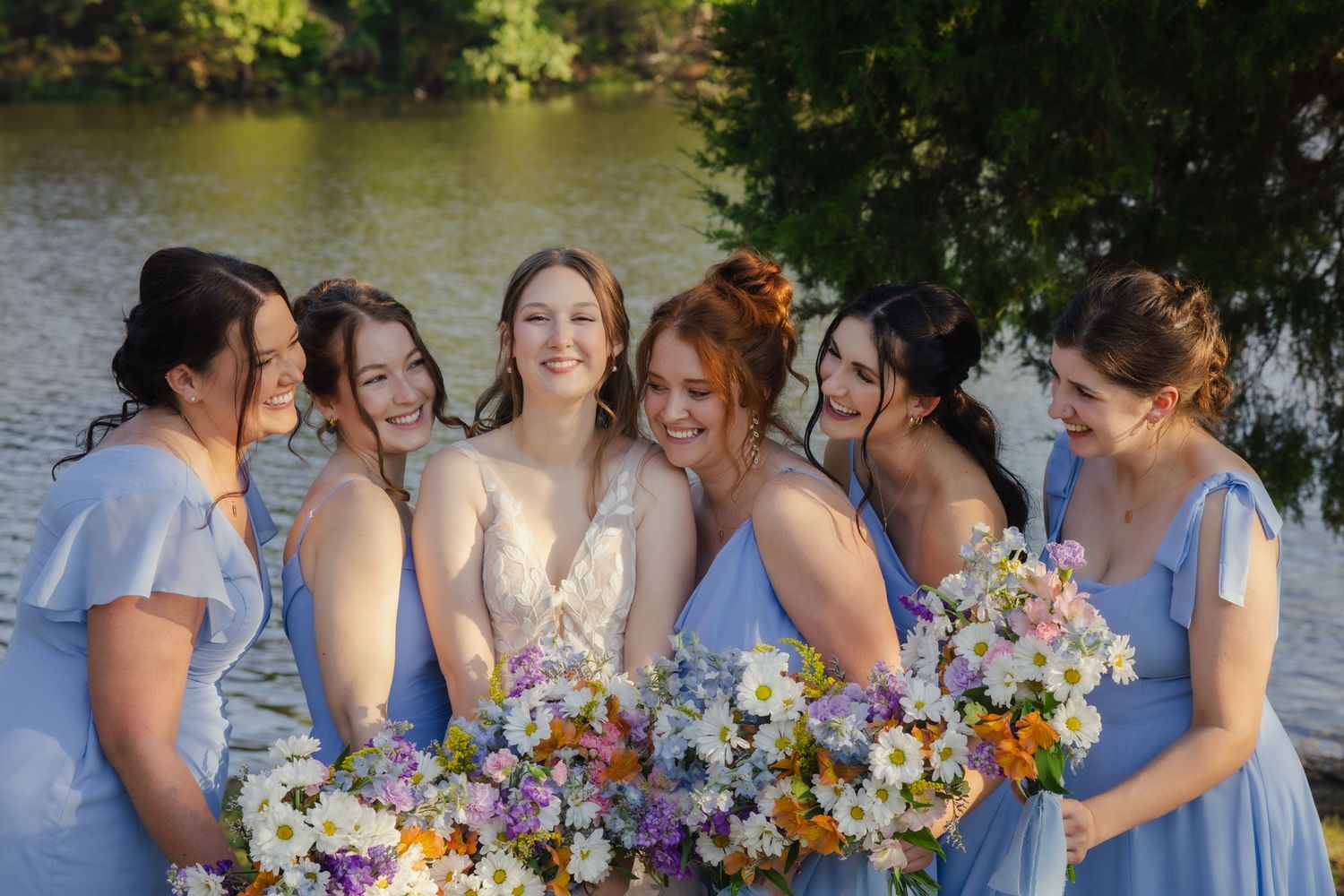 Bridesmaids in light blue dresses holding colorful bouquets pose by lake.