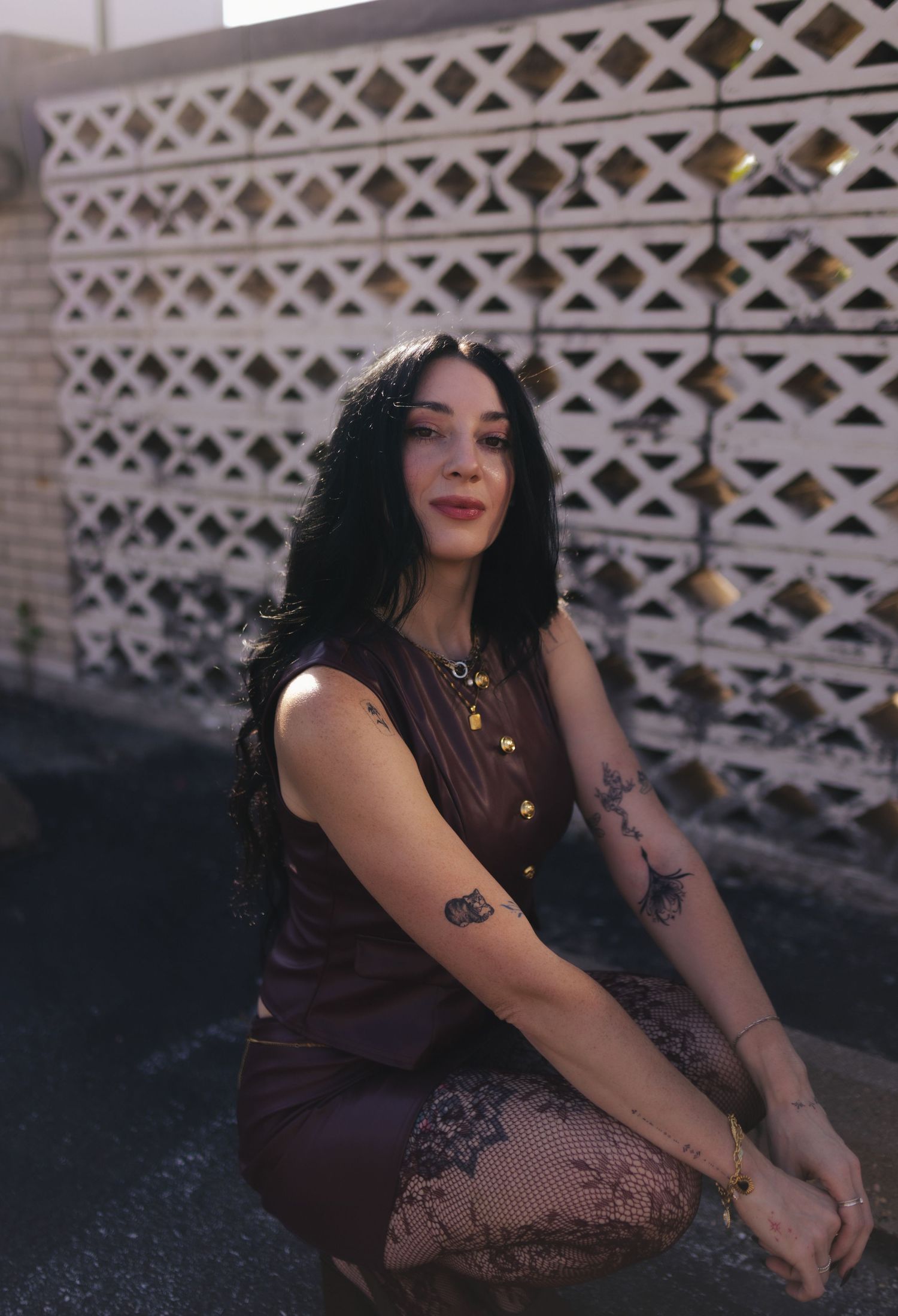 Woman with dark hair and tattoos wearing a brown dress and fishnet tights, posing outdoors near a decorative white wall.
