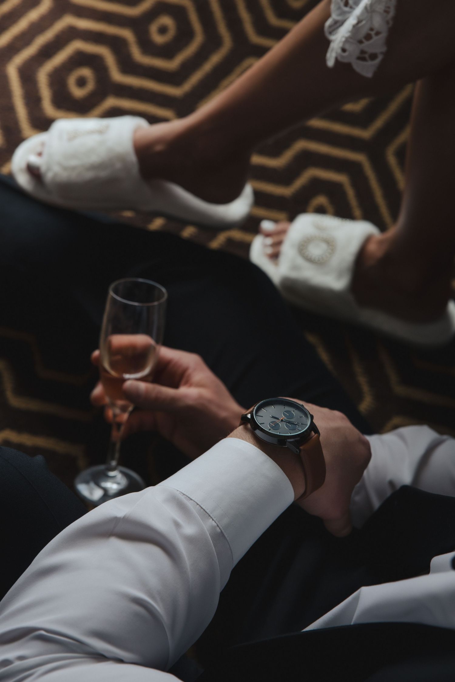 Person in white dress shirt checks watch while holding champagne glass on geometric patterned carpet.