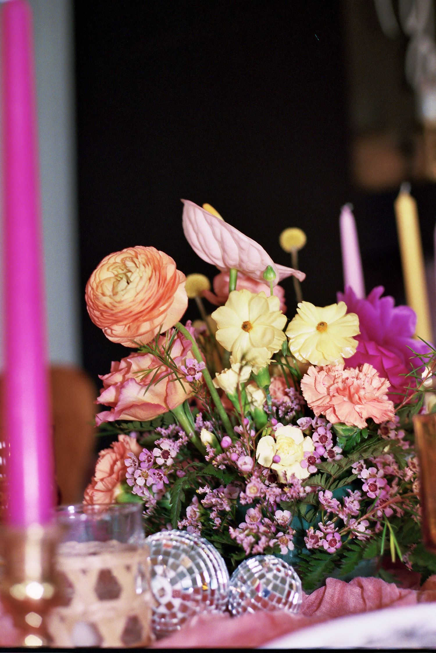 A vintage-style floral arrangement with ranunculus, carnations and purple flowers beside disco balls and tall candles.