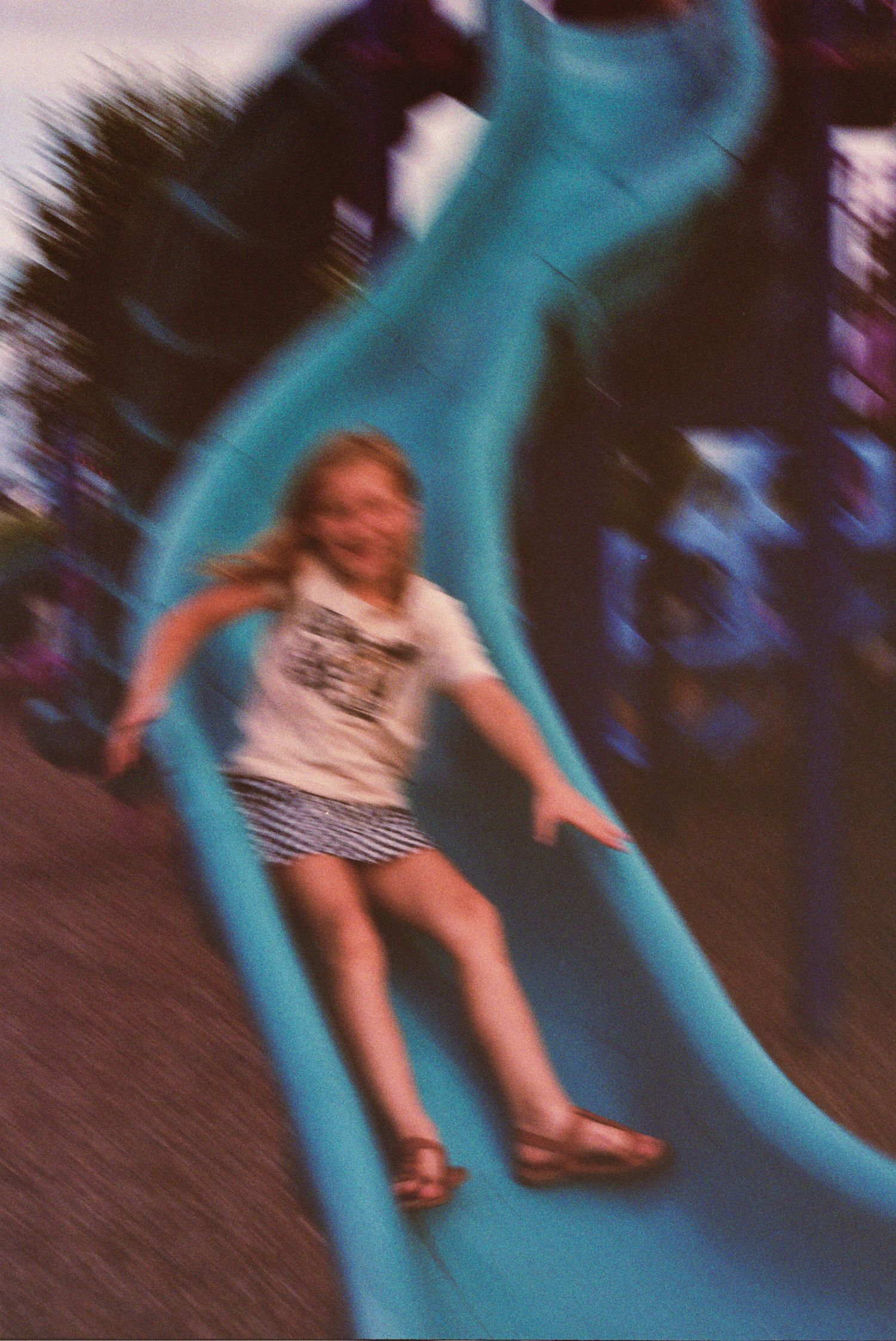 Motion-blurred image of someone sliding down a bright turquoise playground slide with a playful expression.
