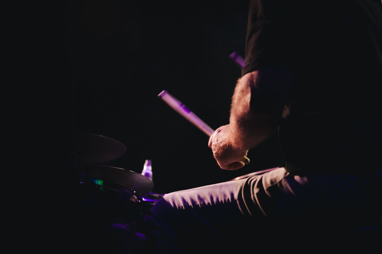 Drummer playing with glowing purple drumstick on stage in dramatic dark lighting during live performance.