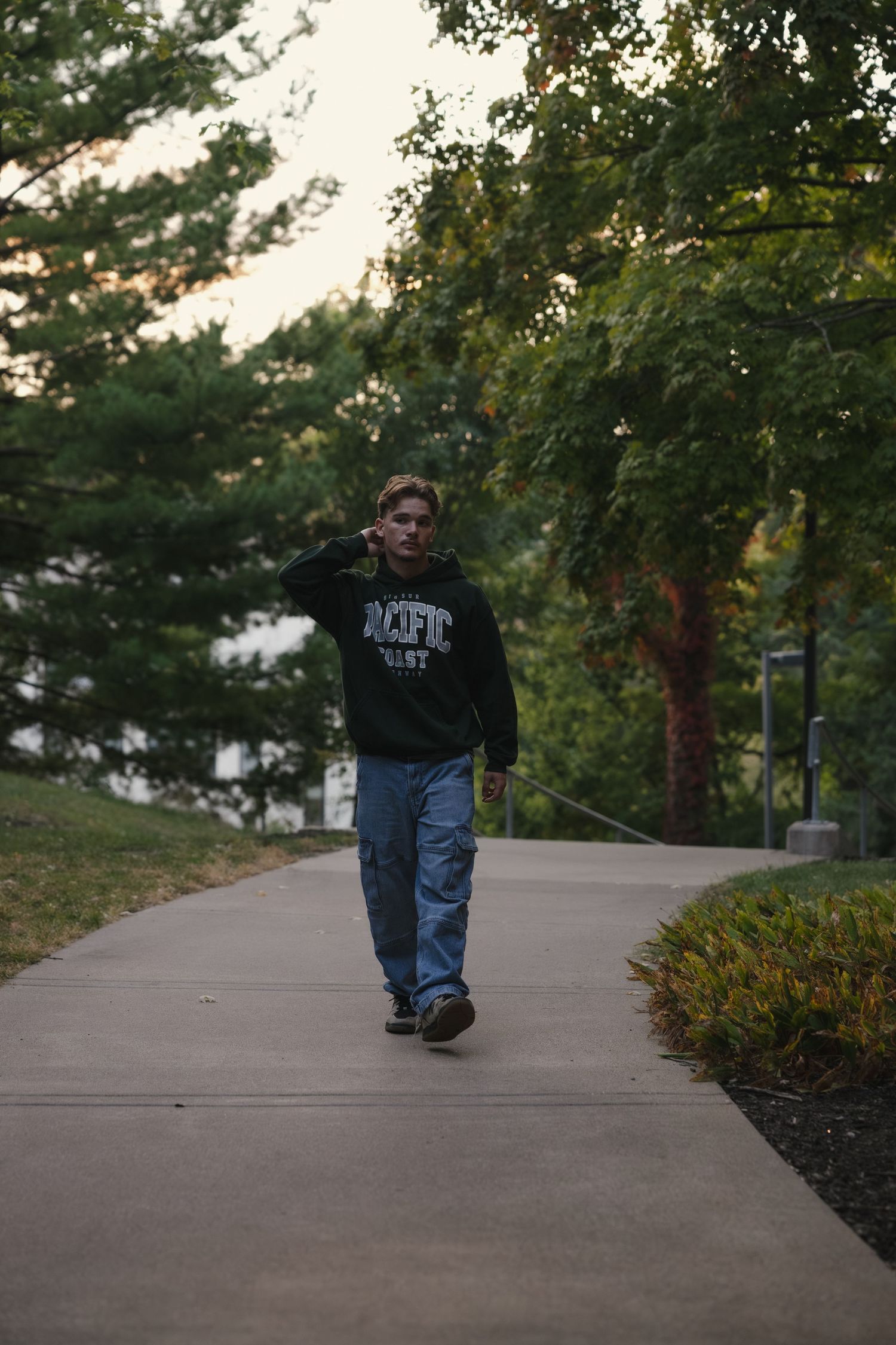 Young man in ROTC hoodie and jeans walking on a park pathway surrounded by green trees.