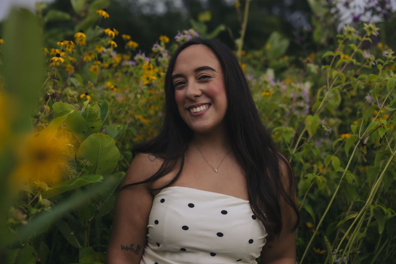 Young woman smiling joyfully among wildflowers and green foliage, wearing a white polka dot top outdoors.
