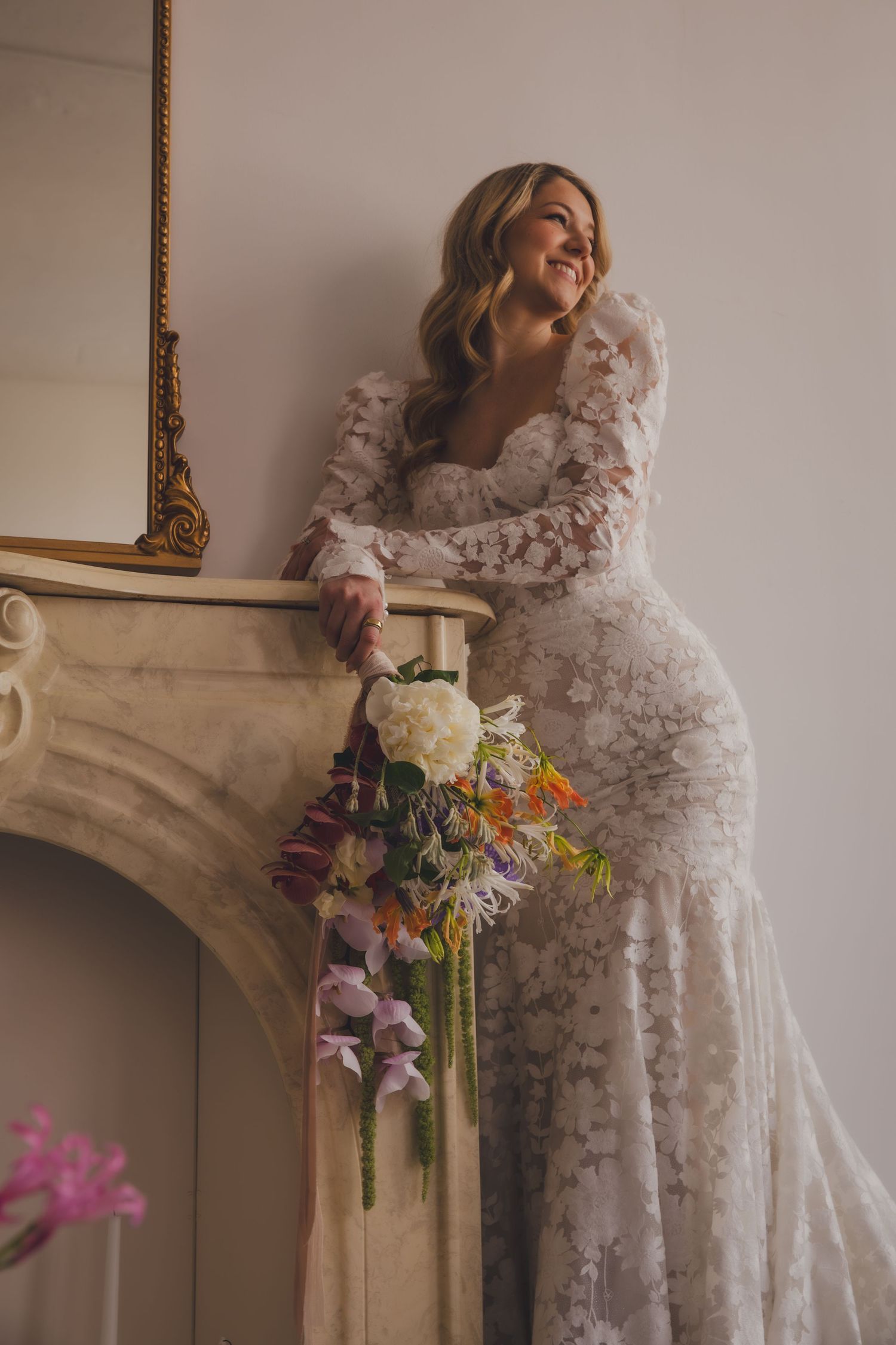 A bride in a long-sleeved lace gown poses by an ornate fireplace with a floral bouquet.