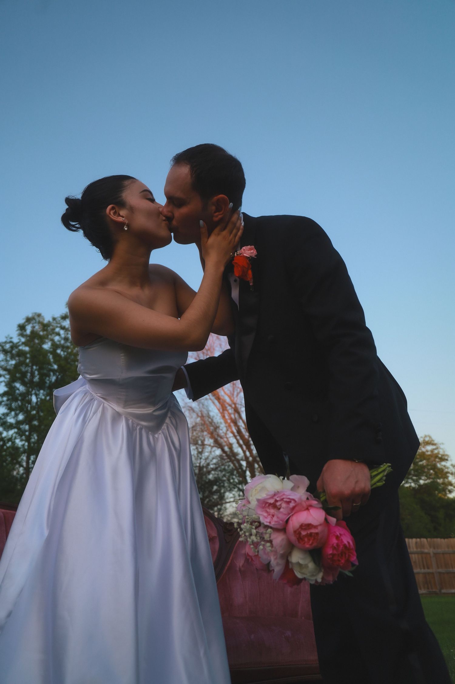 A bride in a white dress and groom in a black suit share a romantic kiss outdoors during their wedding ceremony.