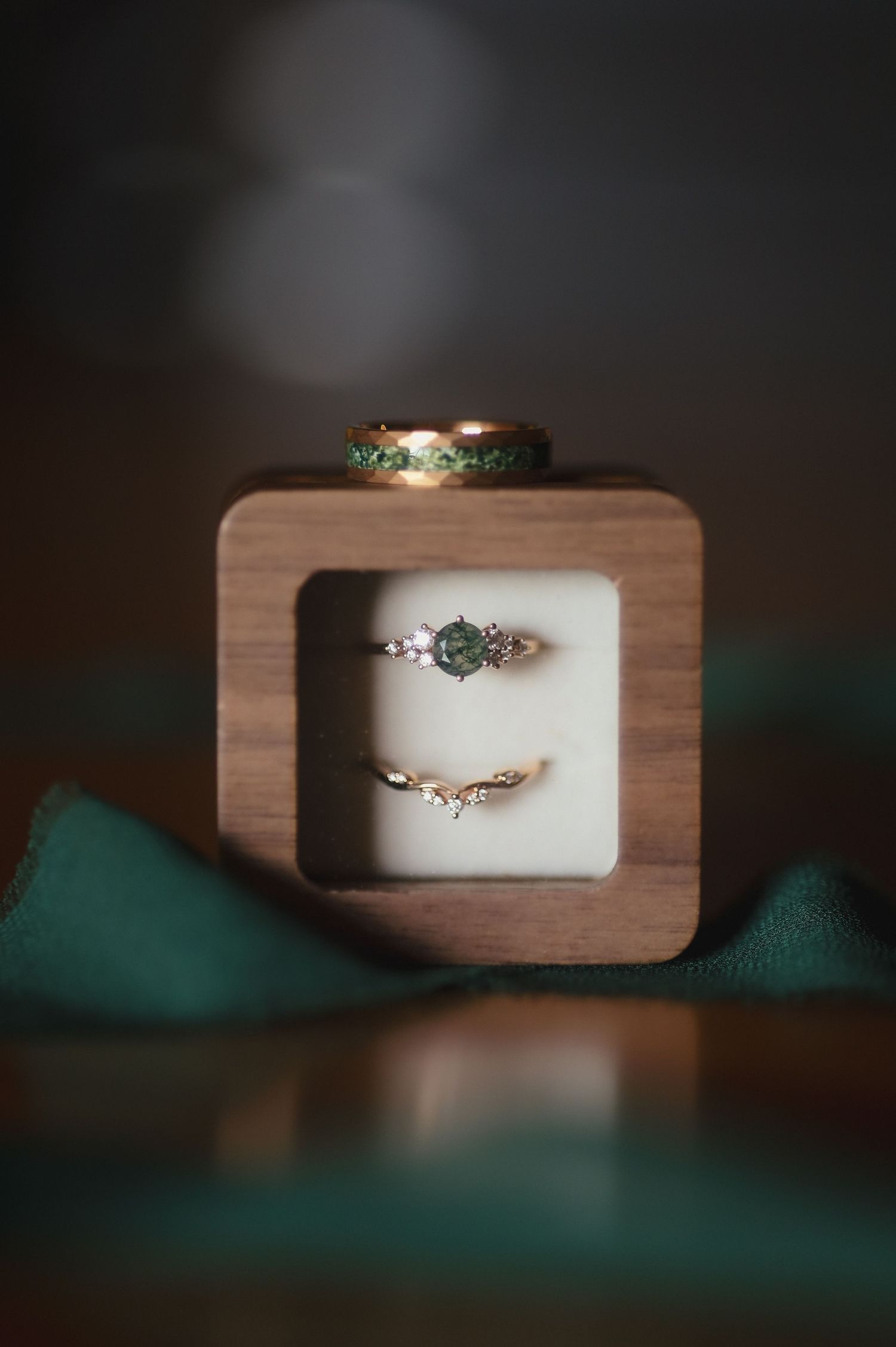 Wedding rings displayed in a wooden box with emerald accents and teal fabric in soft moody lighting.