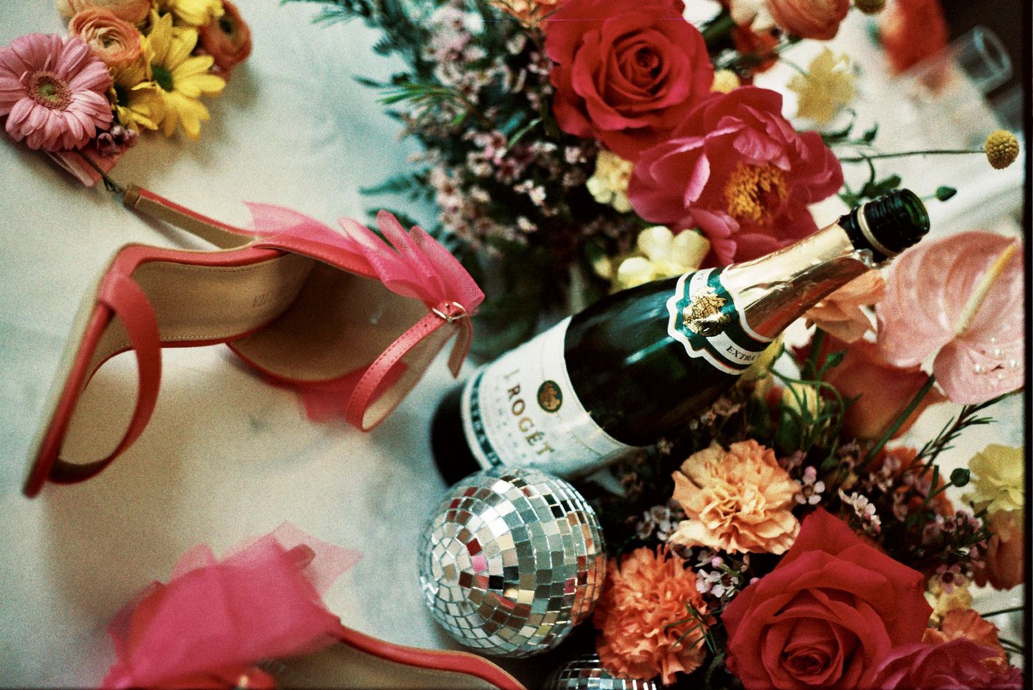 A festive still life with red roses, sunglasses, a champagne bottle and disco ball among colorful flowers.