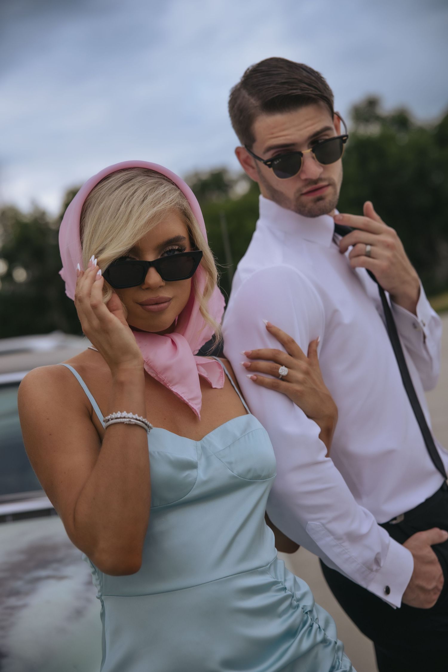 A couple shares a lighthearted moment together outdoors wearing sunglasses and formal attire, with her in mint and him in white.