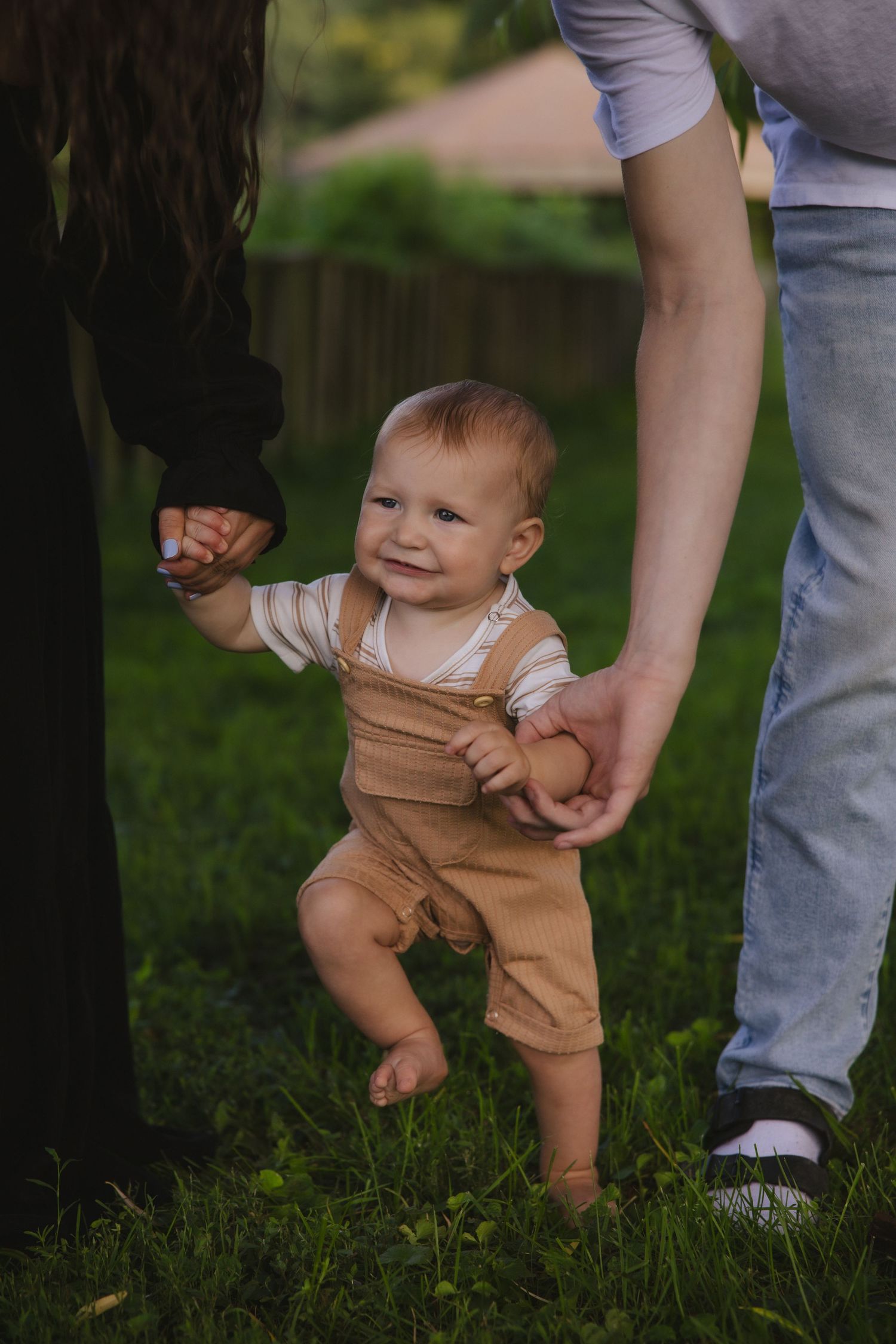 Baby in tan overalls takes first steps while holding onto adult's hands in a grassy outdoor setting.
