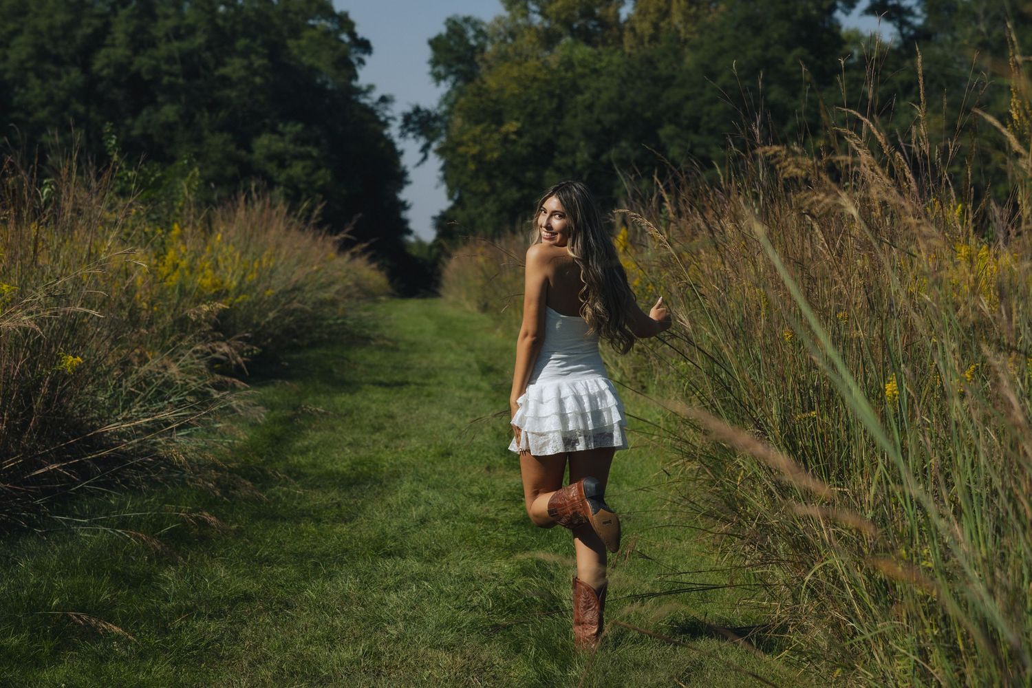 A person walking along a grassy trail through tall wild grass and trees in summertime.