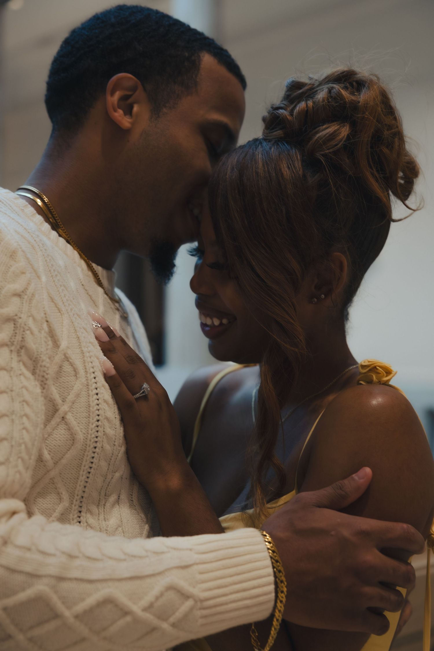 A smiling couple in gold jewelry share an intimate moment, the man in a cream cable-knit sweater embracing a woman in a yellow top.