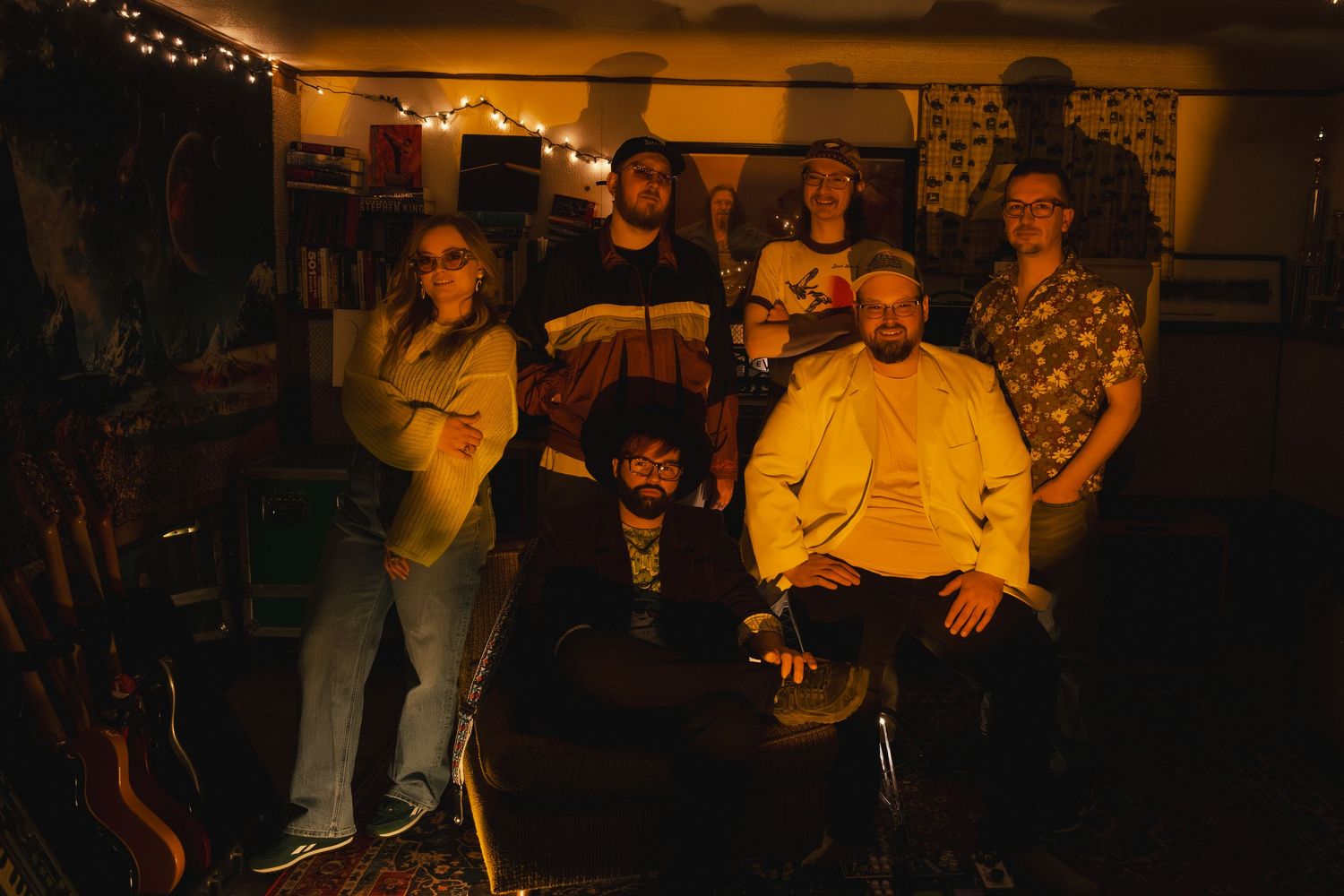 A group of seven musicians posed together in a warmly lit room with string lights and musical equipment.