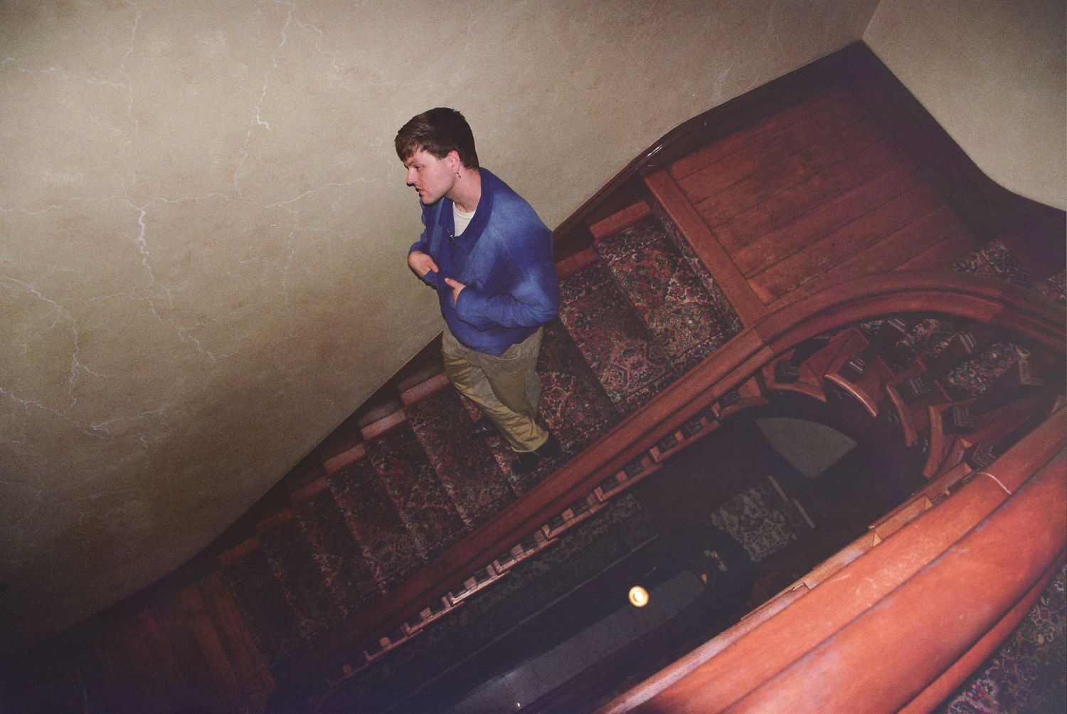 Man in blue sweater descending ornate wooden spiral staircase in dimly lit historic building.