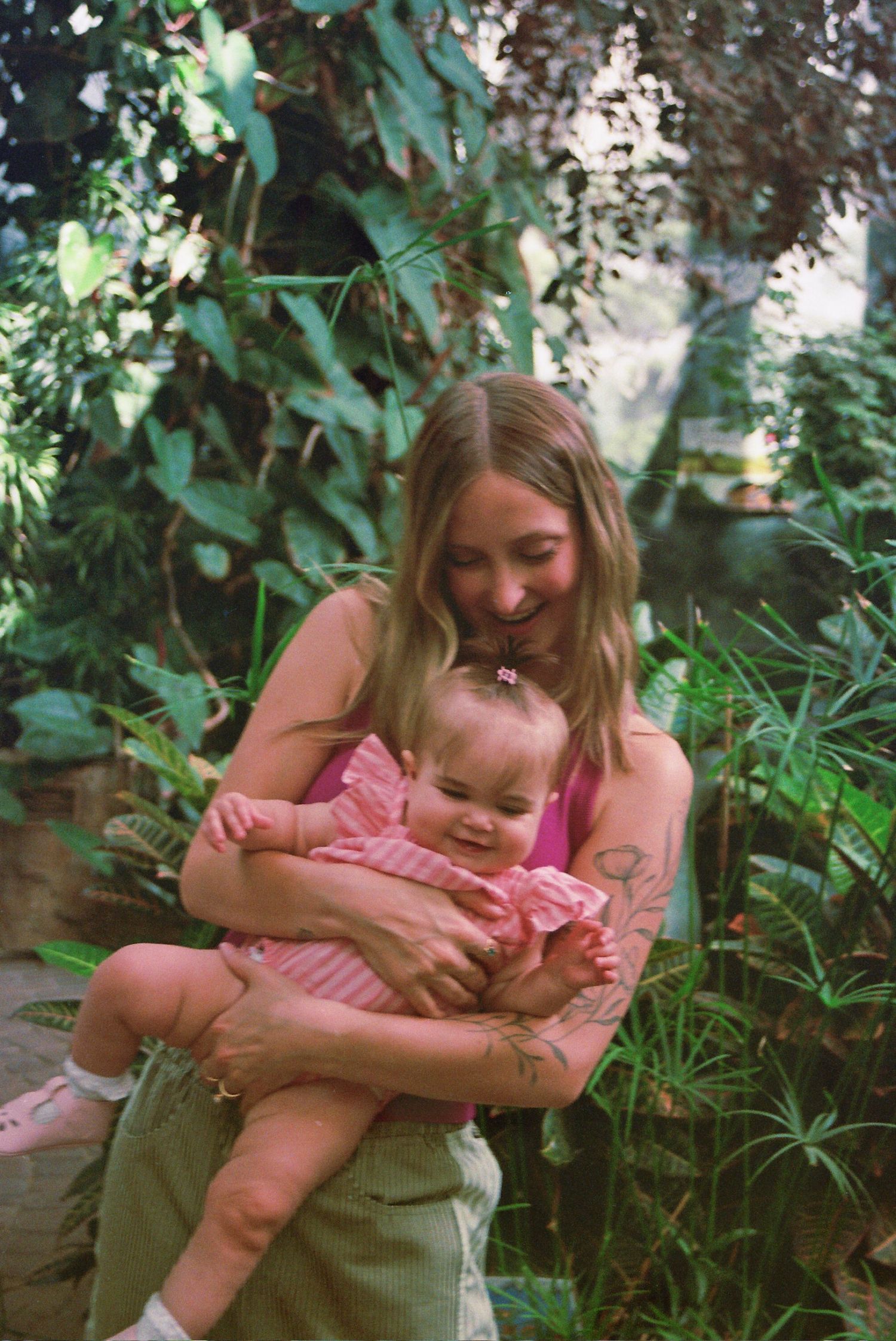 A young woman with blonde hair holds a smiling baby in pink outdoors among lush green tropical plants.