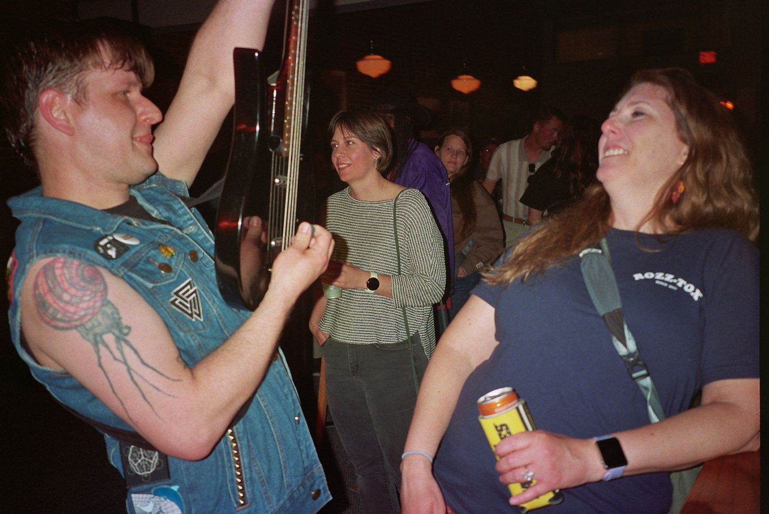 Two people dancing and laughing at a lively bar event, with a tattooed man in a vest and a woman holding a drink.