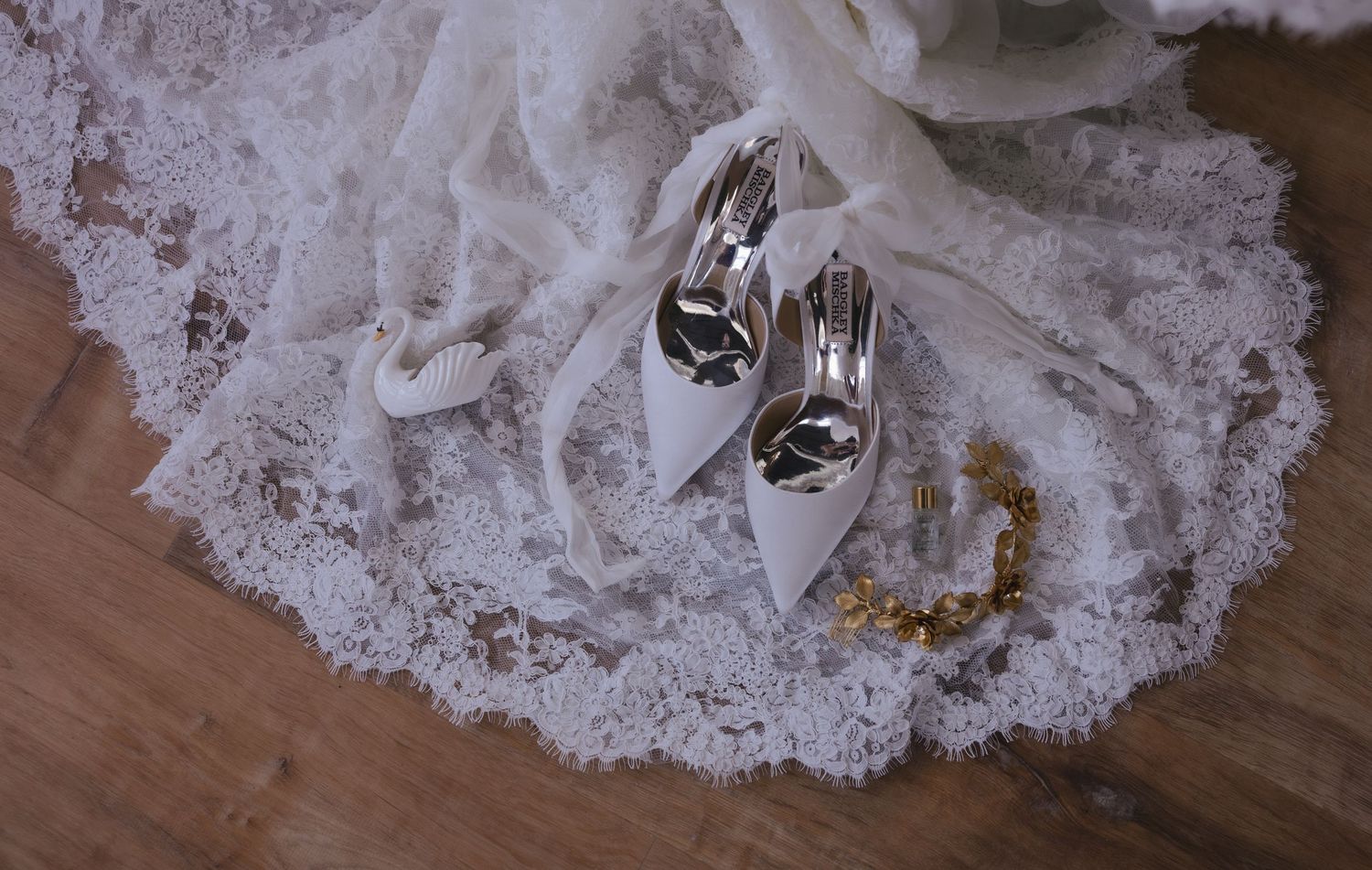 White bridal heels resting on delicate lace wedding veil with gold accents on wooden floor.