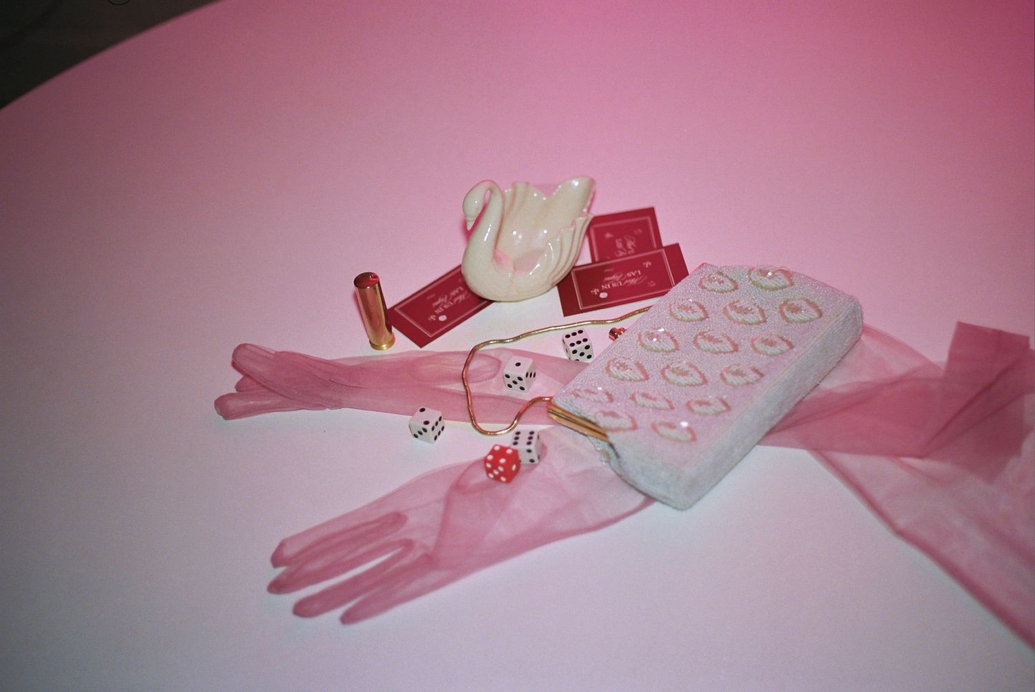 Pink aesthetic flat lay with heart-embellished clutch, long gloves, swan figurine, dice, and cosmetics on pink background.