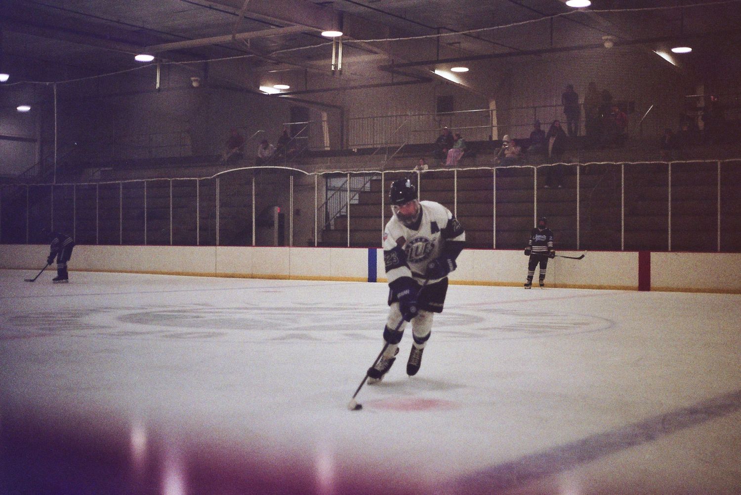 Hockey player skating with puck on indoor ice rink during game, spectators visible in background bleachers.