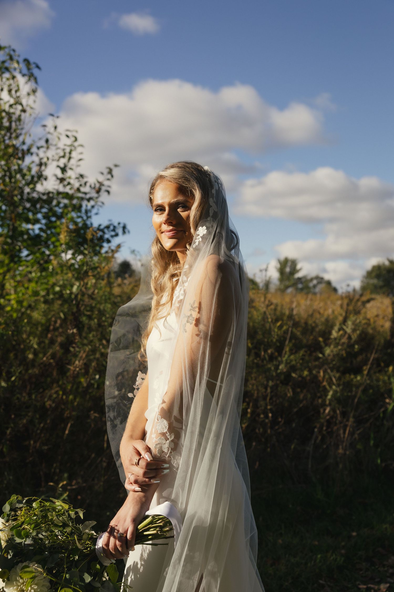 A bride in a flowing white veil stands in a garden setting against a blue sky with puffy clouds.