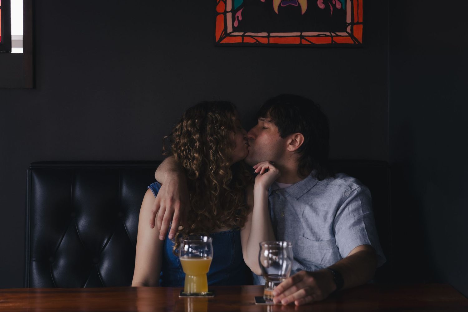 Couple kissing romantically at a bar table with glasses of beer and water in a dimly lit restaurant.