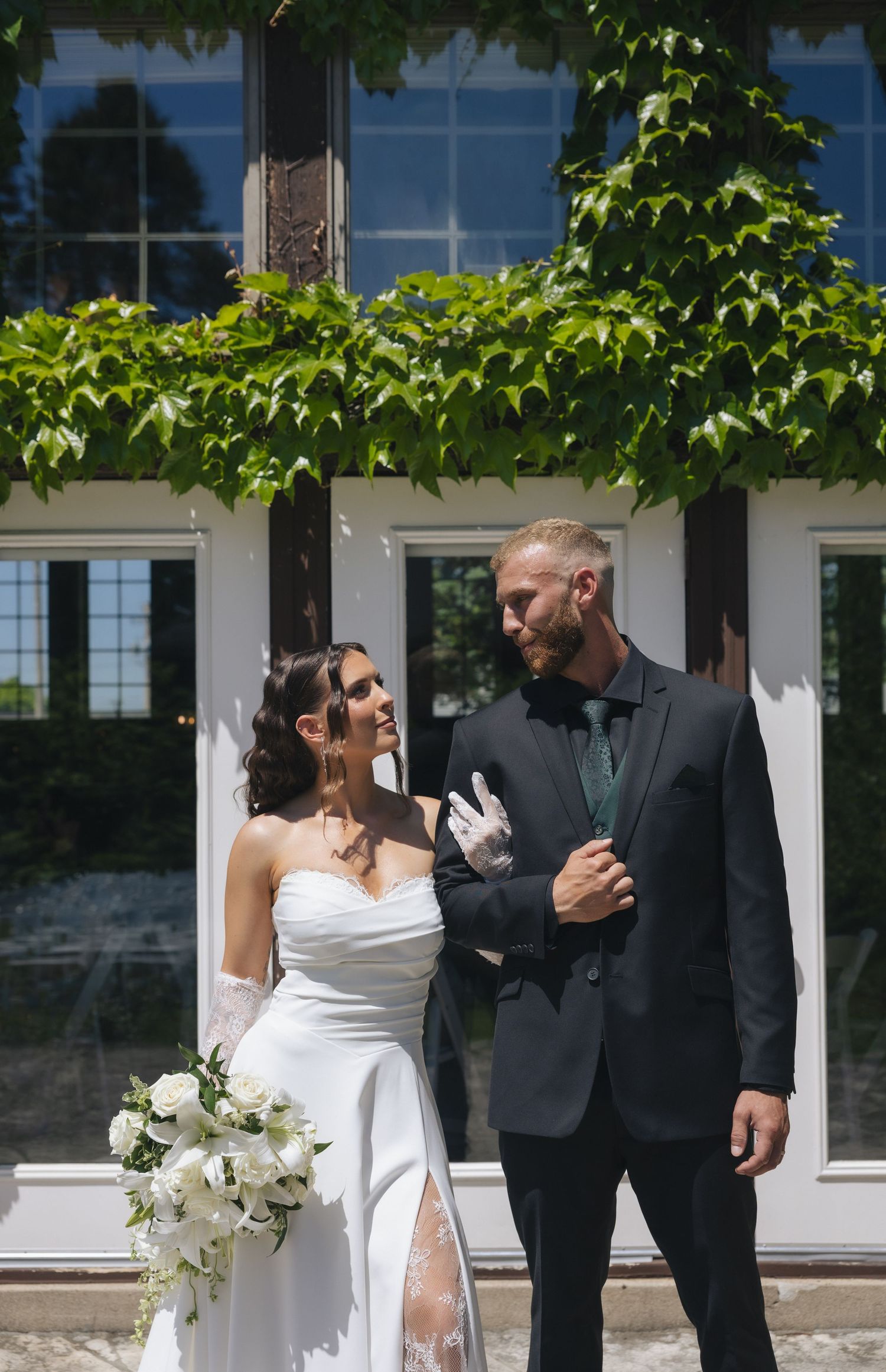 Bride in white gown holding bouquet stands with groom in dark suit outdoors before ivy-covered building on wedding day.