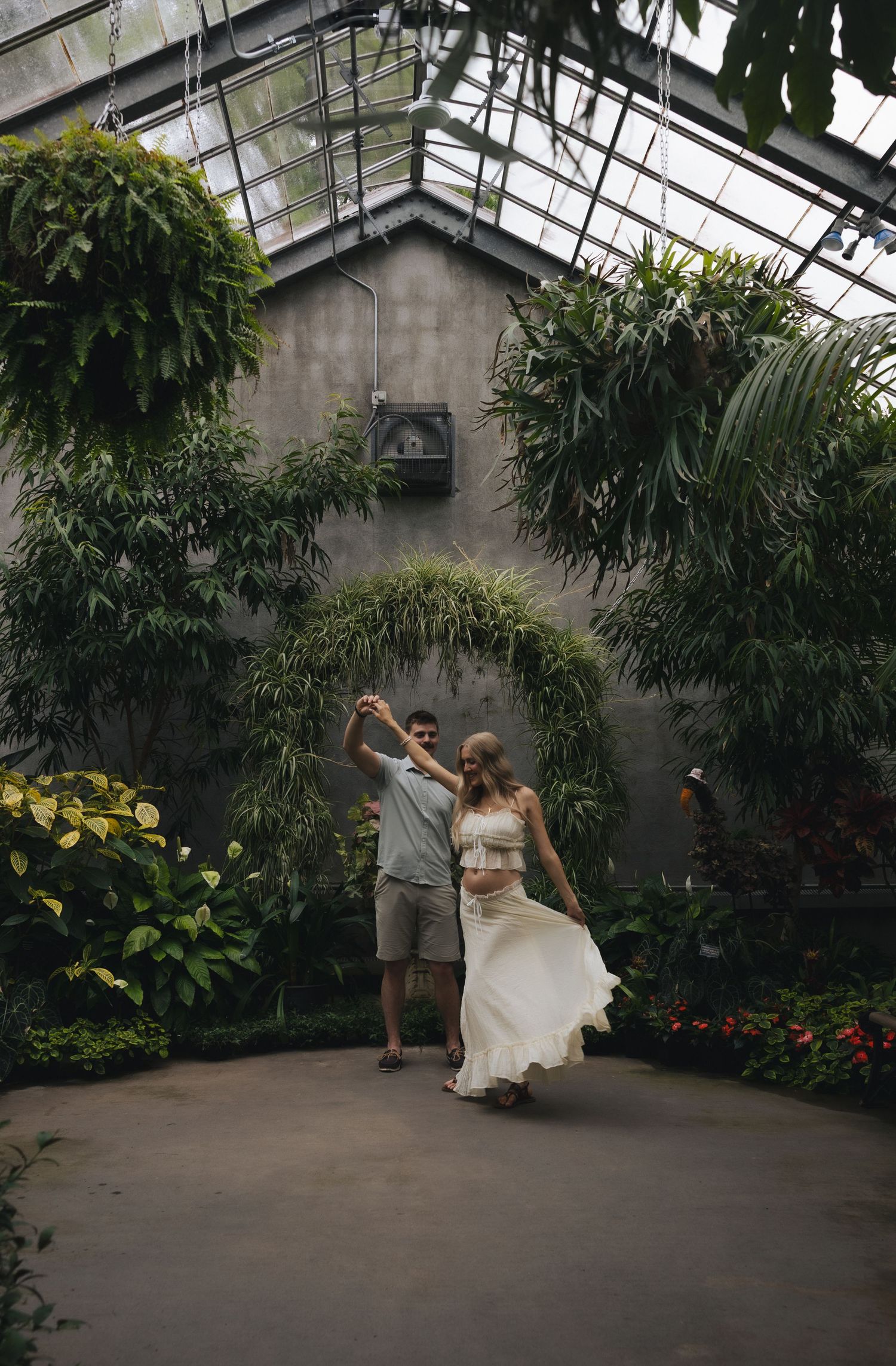 Couple dancing joyfully in a lush greenhouse conservatory surrounded by tropical plants and natural light.