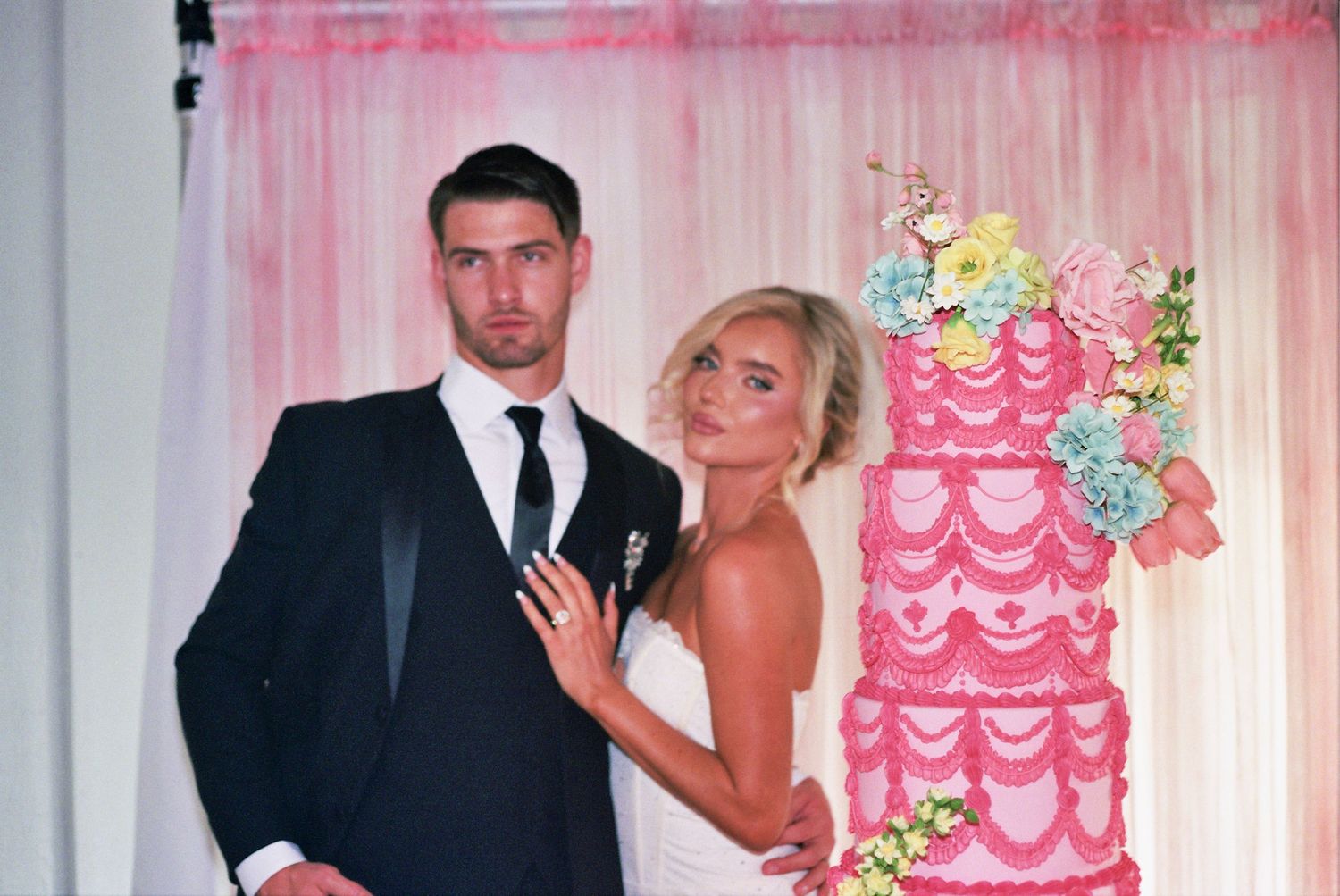People celebrate at a wedding reception with a pink and white tiered cake and floral decorations in the background.