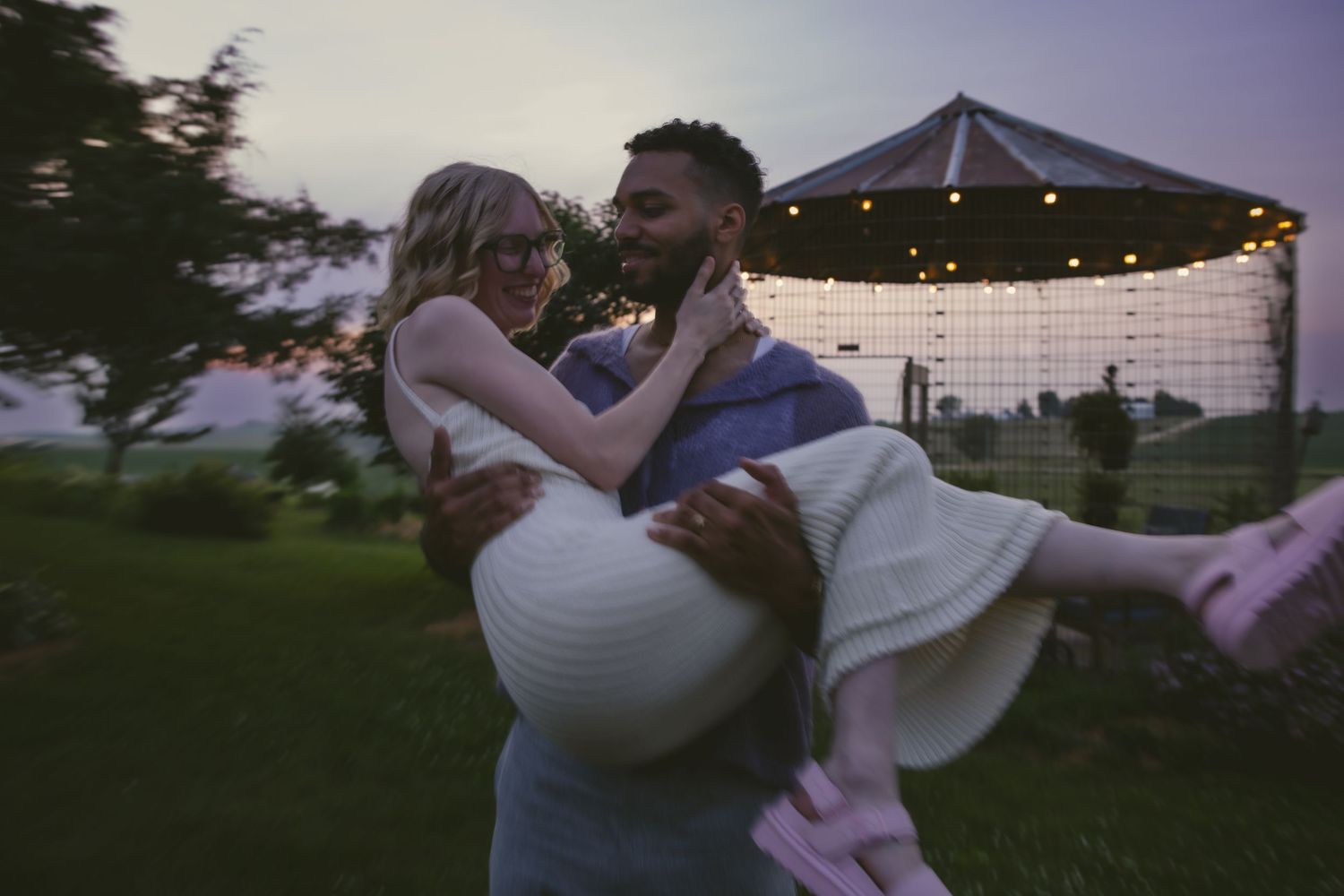A joyful couple embraces during twilight beneath a romantic gazebo decorated with string lights.