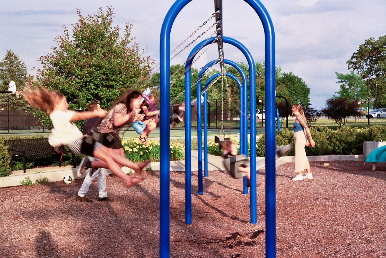 People playing on a row of blue metal swings at a playground with rubber mulch ground covering.