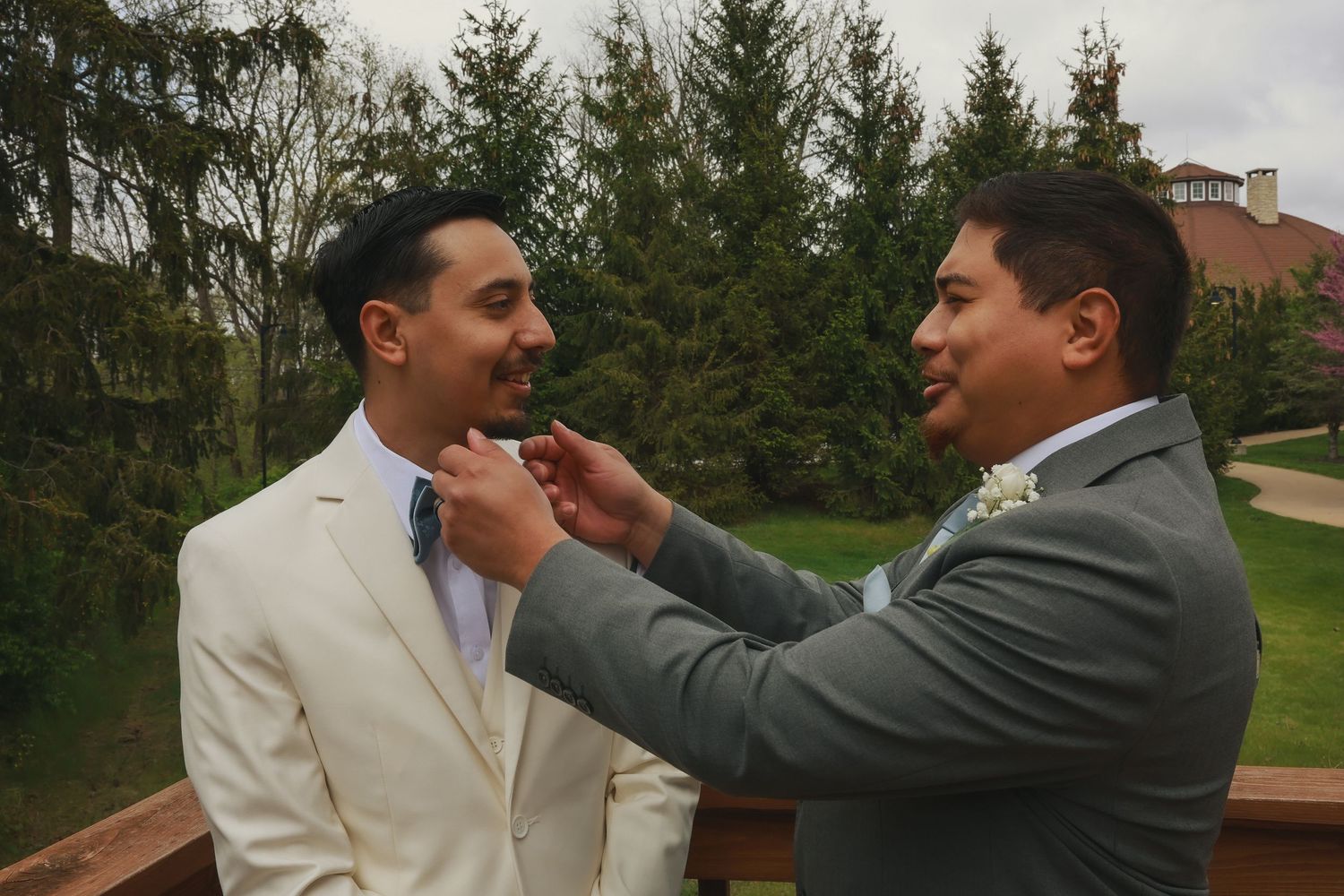 Two people in formal attire stand outdoors as one adjusts the other's tie against a backdrop of evergreen trees.